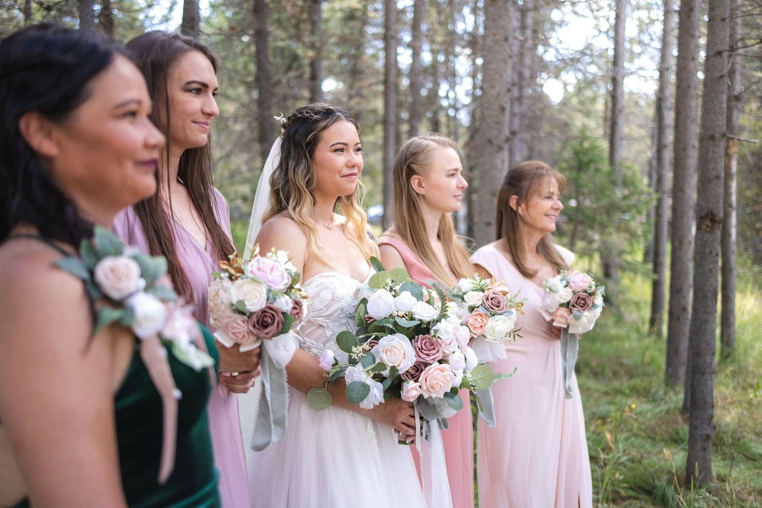 Bridal party poses with bouquets in wooded outdoor setting wearing pastel dresses.