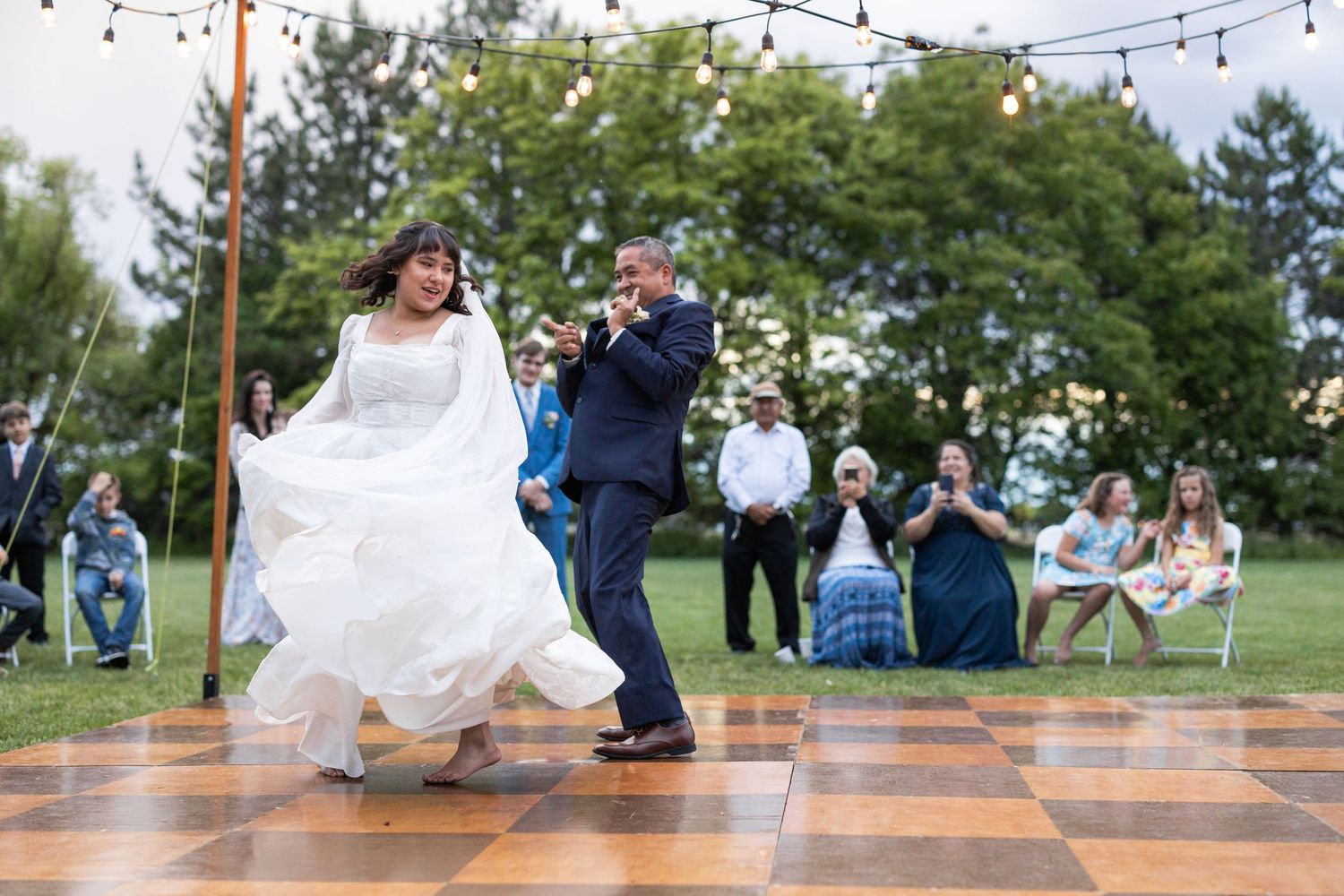 A flowing white wedding dress twirls during a dance on an outdoor wooden floor.