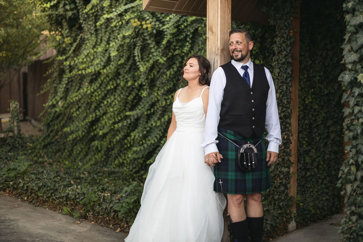 Wedding couple stands together, groom wearing traditional Scottish kilt beside bride in white dress.