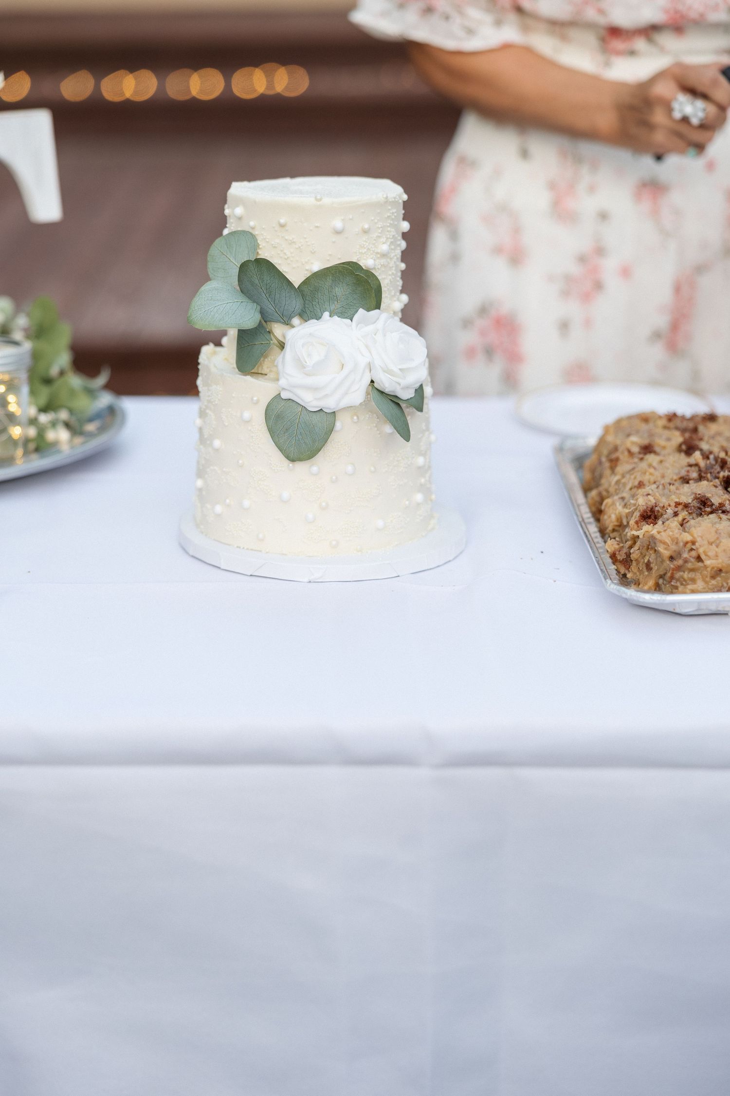 White wedding cake with eucalyptus leaves and roses displayed on white tablecloth.