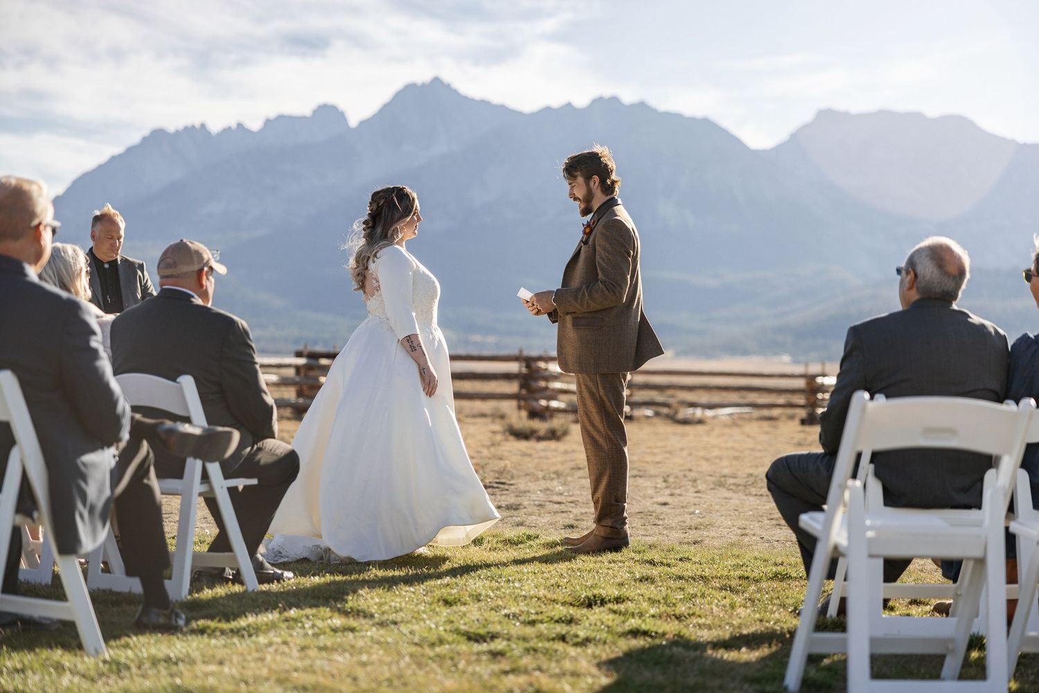 Outdoor wedding ceremony with mountain backdrop and white chairs on grassy hillside.