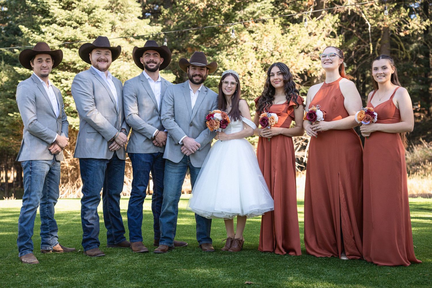 Wedding party in rustic country style with groomsmen in light blue jackets and cowboy hats alongside bridesmaids in rust dresses.