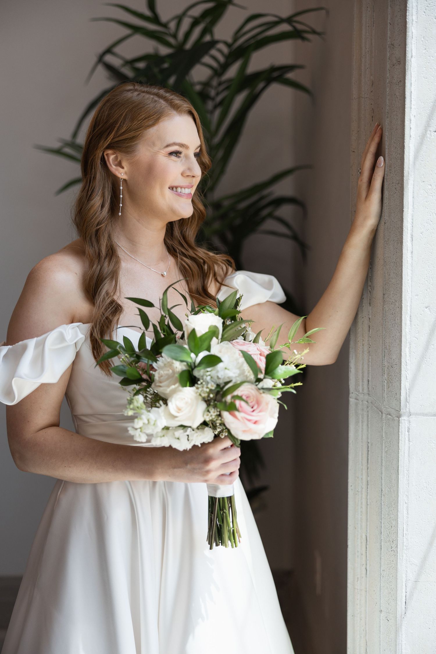 A bride in white holds a beautiful bouquet of roses and greenery while posing against a wall.