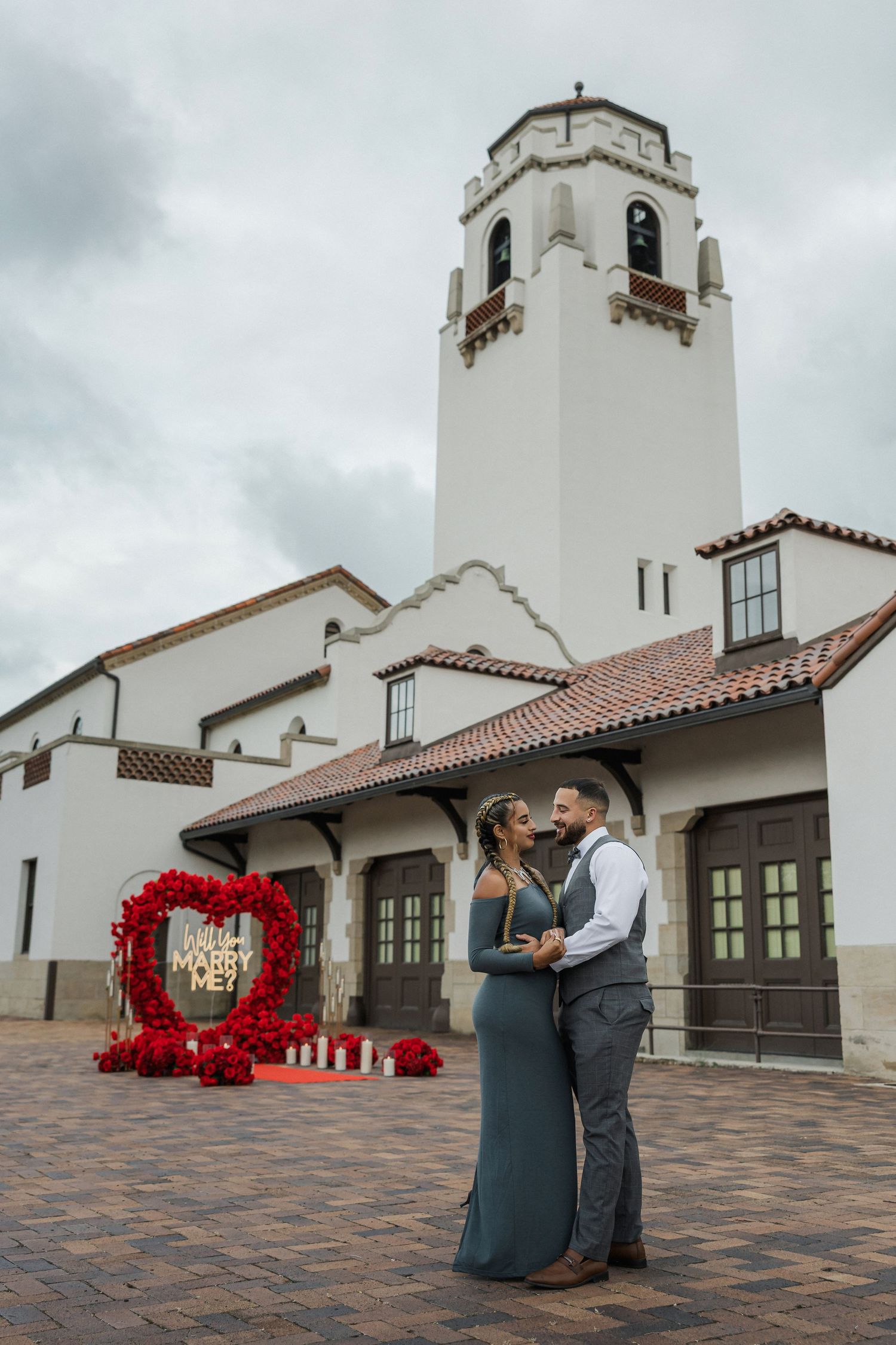 Couple embraces in front of a white Spanish-style building with a red heart floral proposal backdrop on brick plaza.