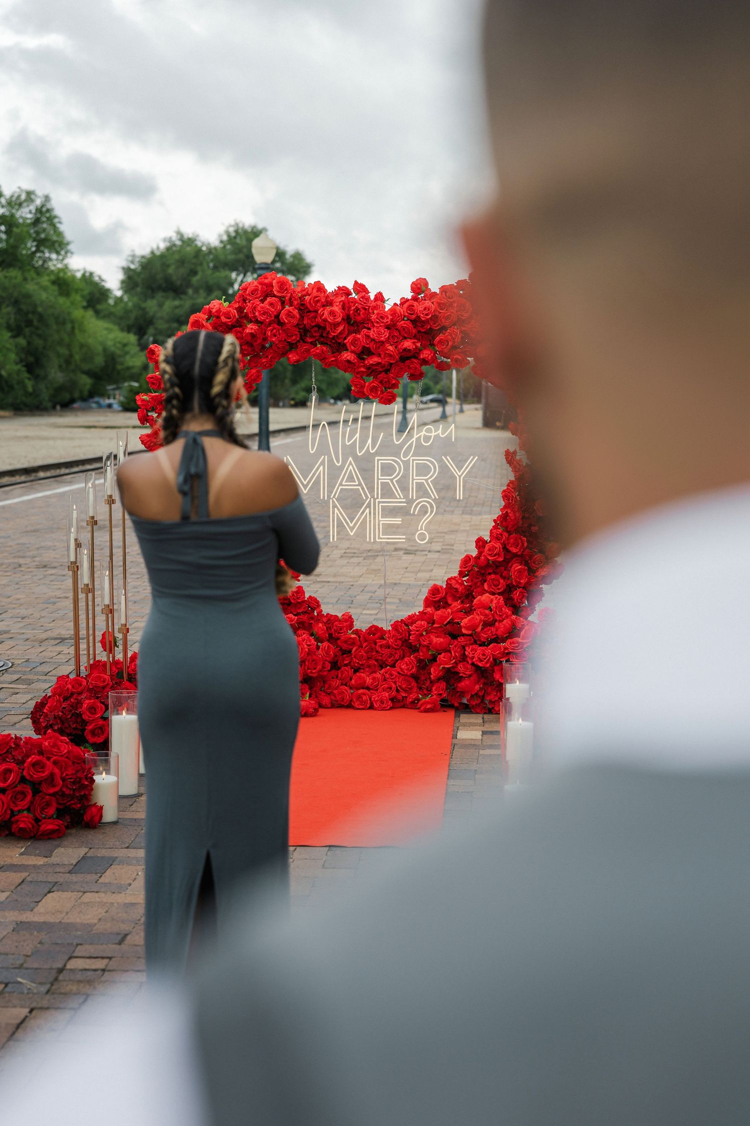 Woman in gray dress stands before a red rose heart arch with Will You Marry Me sign on a brick path.