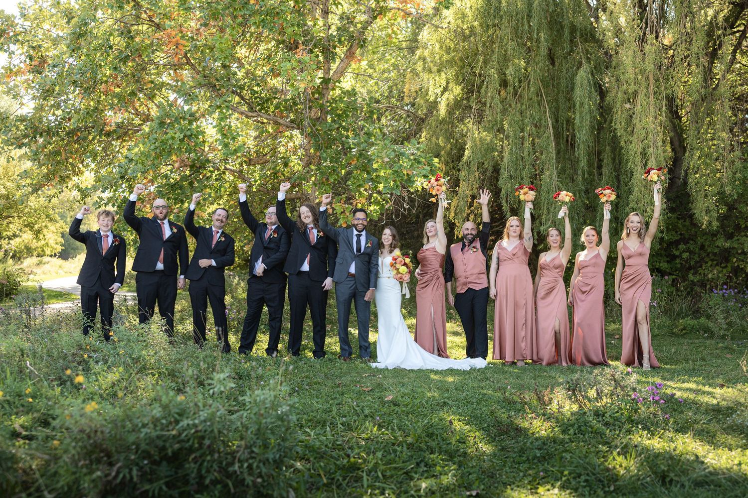 Wedding party in black suits and dusty rose dresses poses under weeping willow trees.