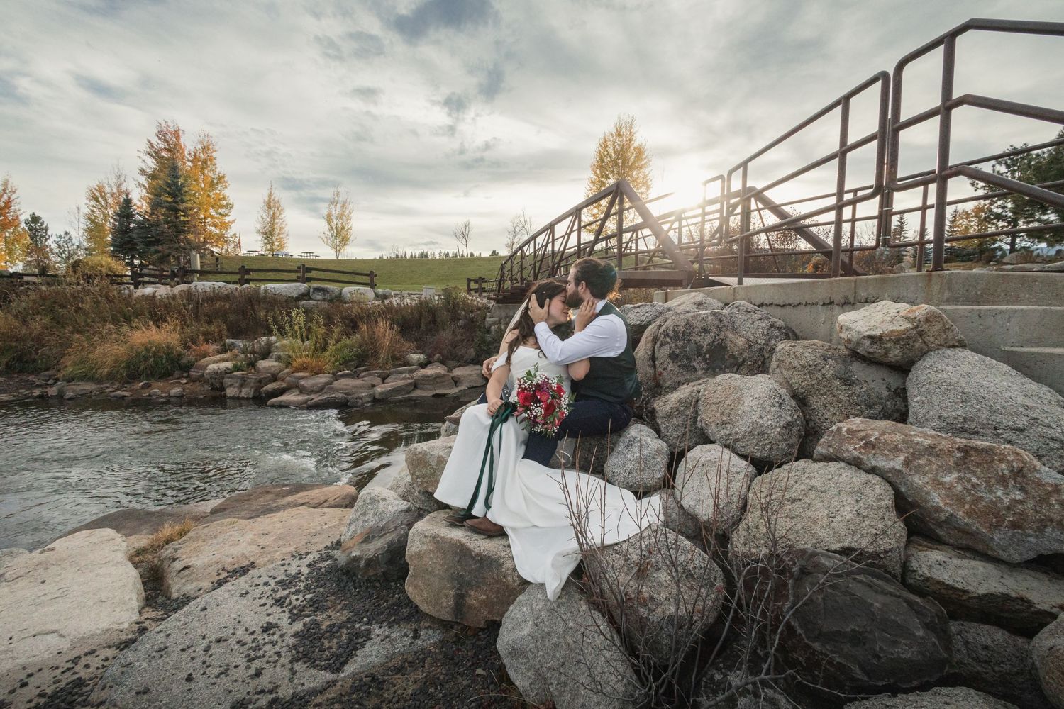 Couple in wedding attire embracing on rocky riverbank beneath metal bridge.
