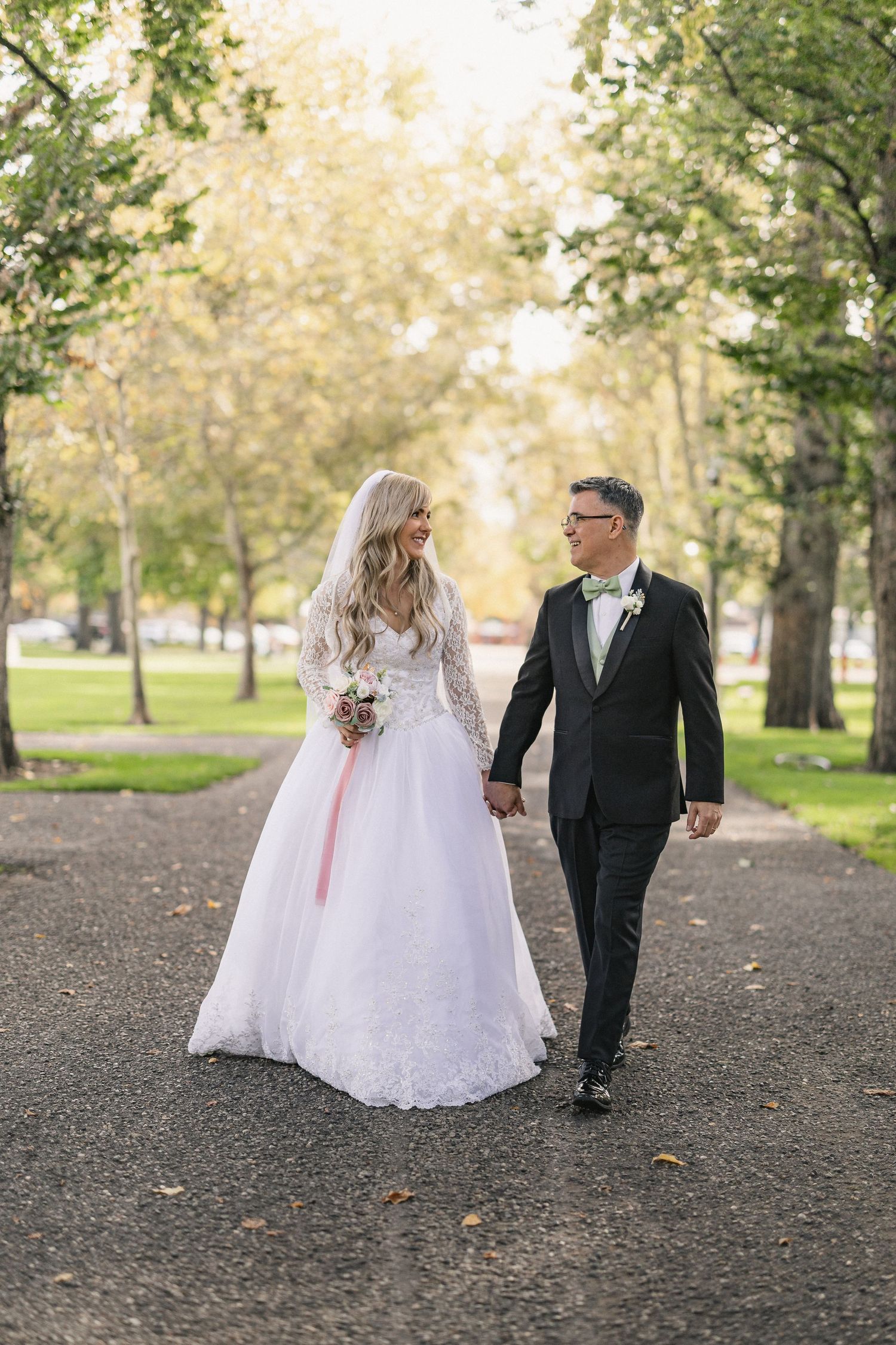 Newlyweds walking hand in hand on autumn path surrounded by trees.