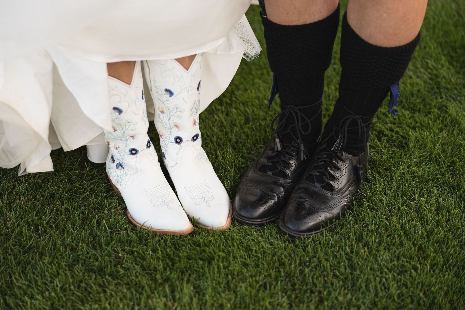 A close-up shot of white bridal shoes and black dress shoes on grass.