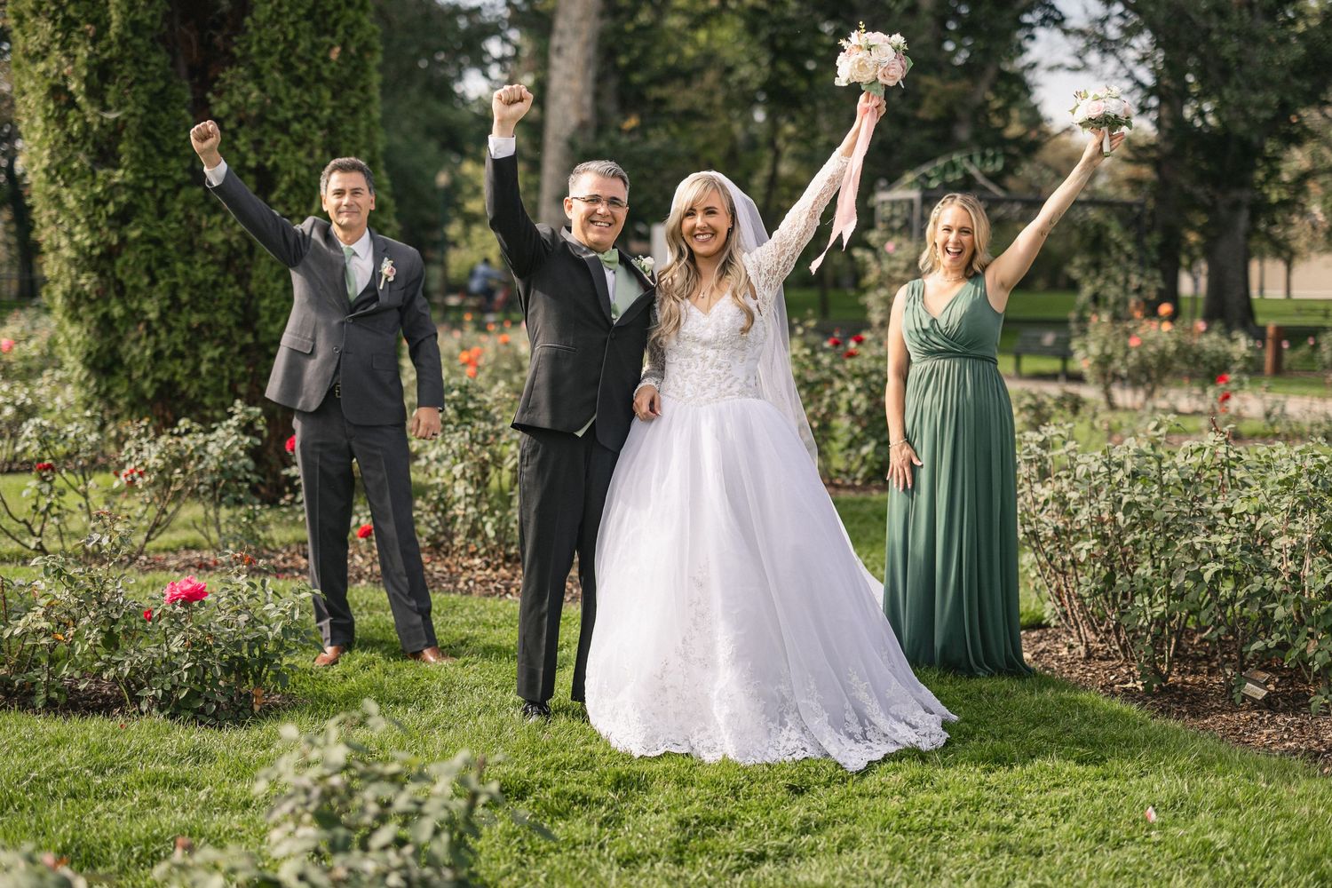 Wedding party celebrates with raised arms and joyful expressions in a garden setting.
