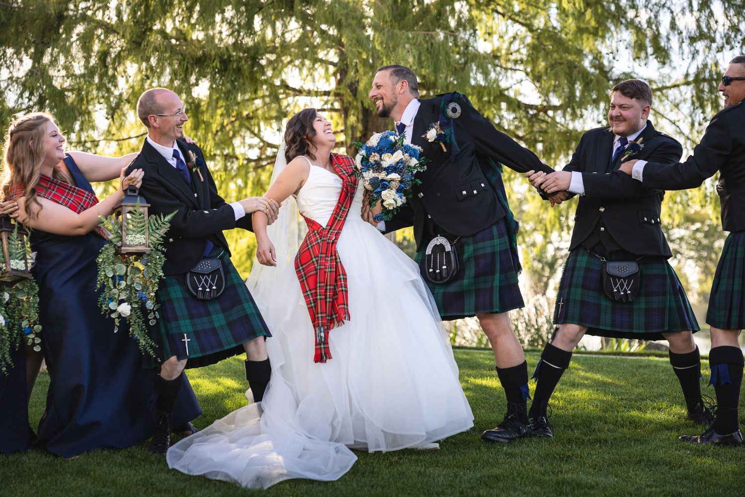 Wedding party celebrates joyfully in Scottish attire with traditional kilts against a backdrop of evergreen trees.