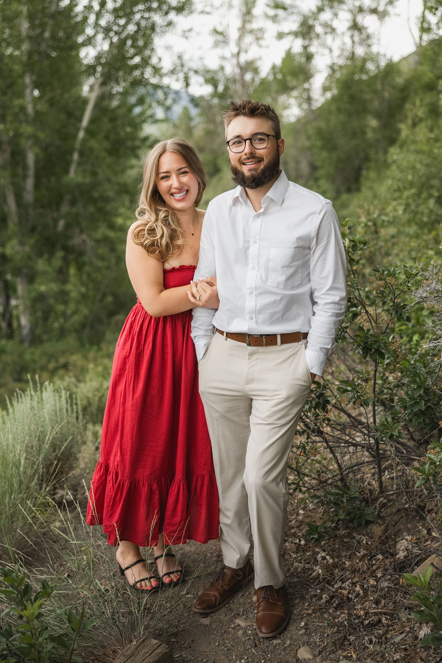 Couple poses together outdoors, with a red dress and white shirt against natural greenery in a romantic photoshoot setting.