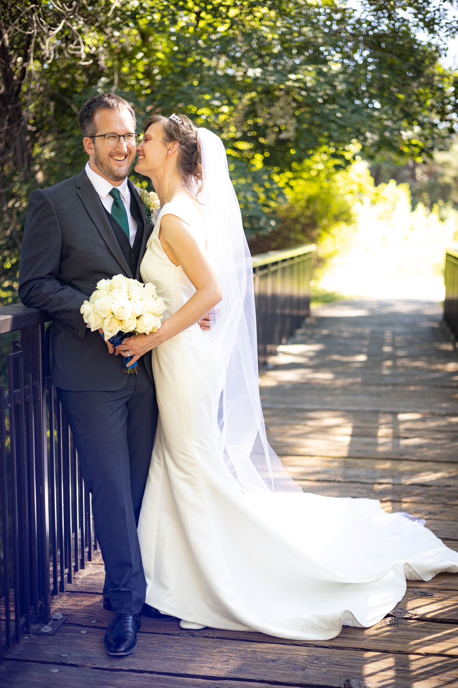 A couple in wedding attire poses on a sunlit wooden bridge.