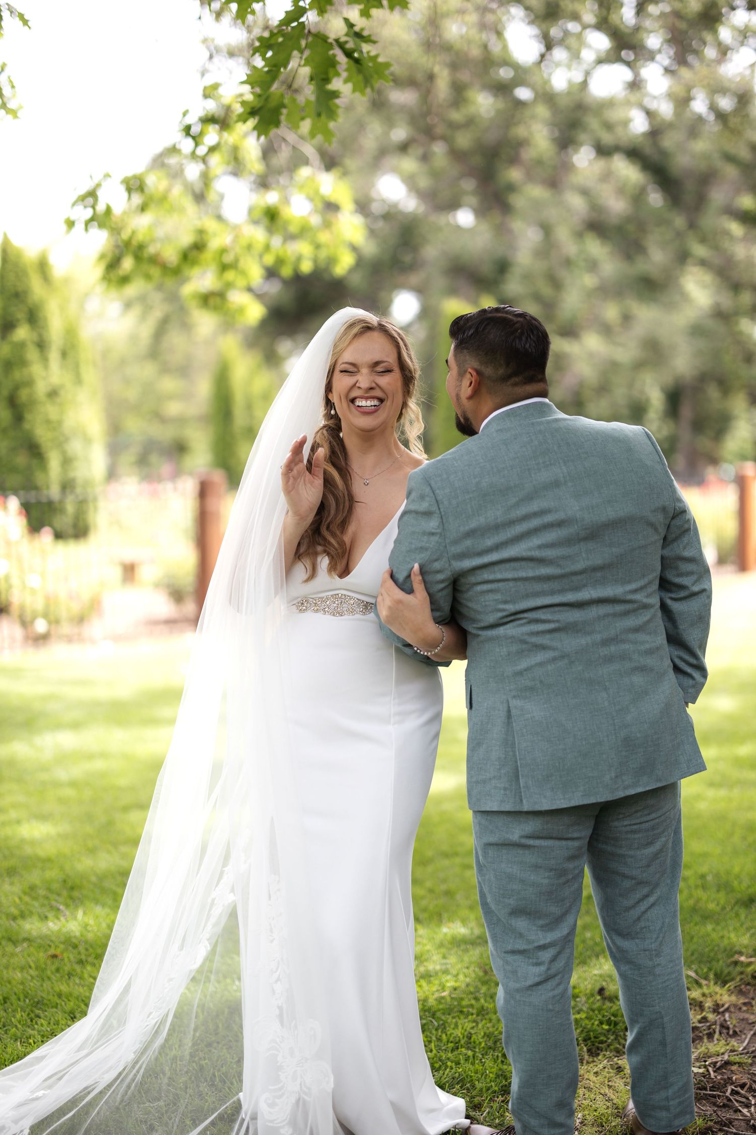 A joyful wedding couple shares a candid moment outdoors with the bride in white and groom in sage green suit.