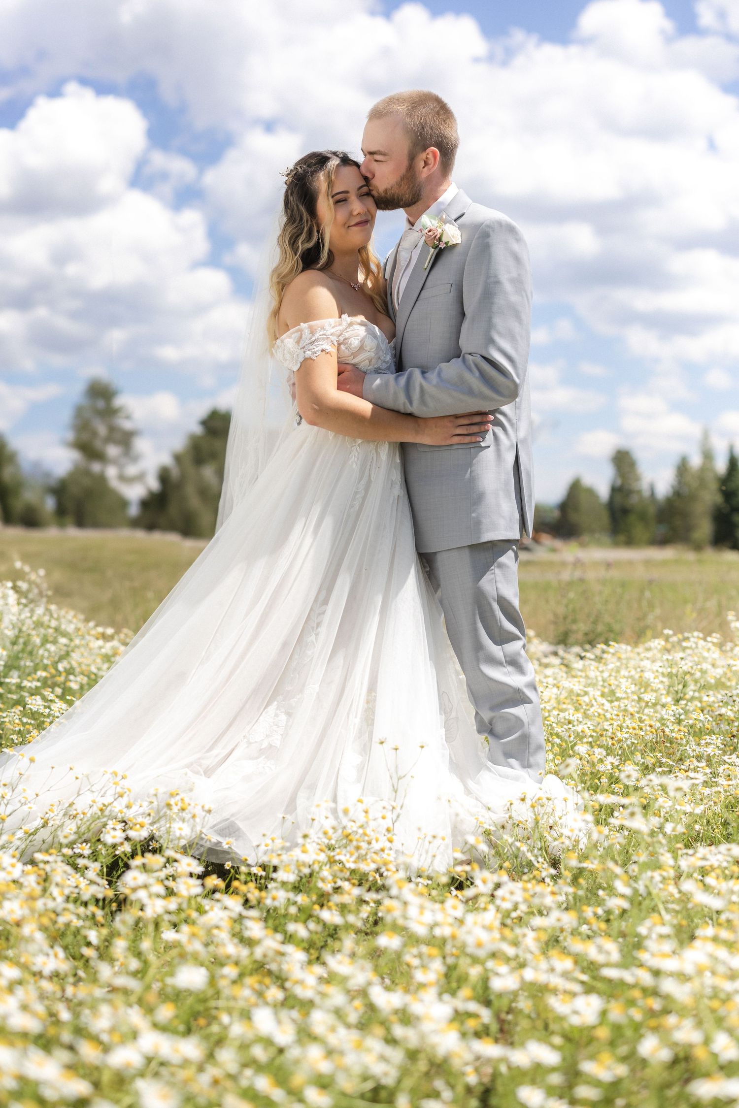 Newlyweds embrace in a meadow of white wildflowers under blue summer sky.