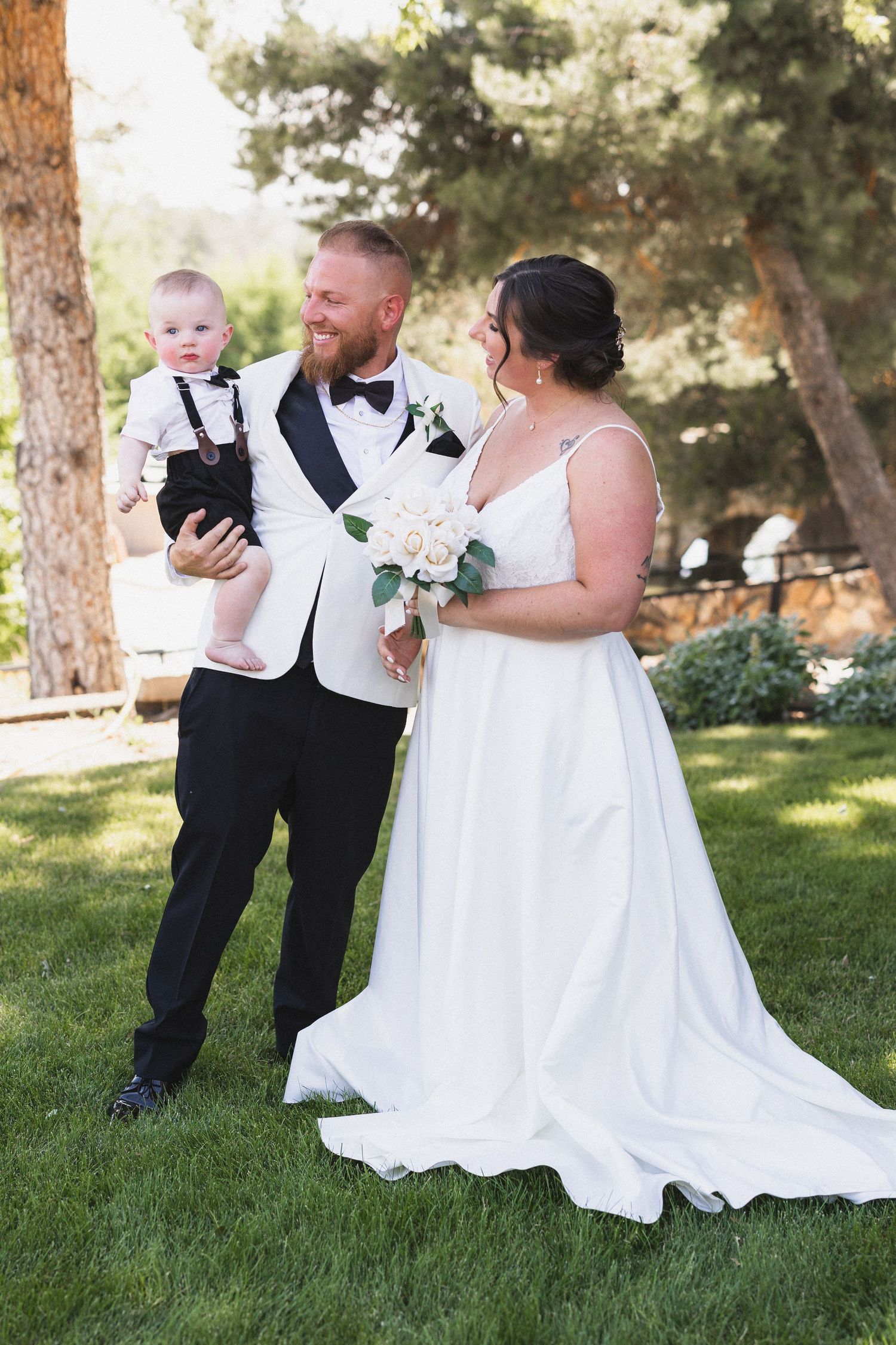 Wedding party poses outdoors with white and black formal attire against a backdrop of tall pine trees.