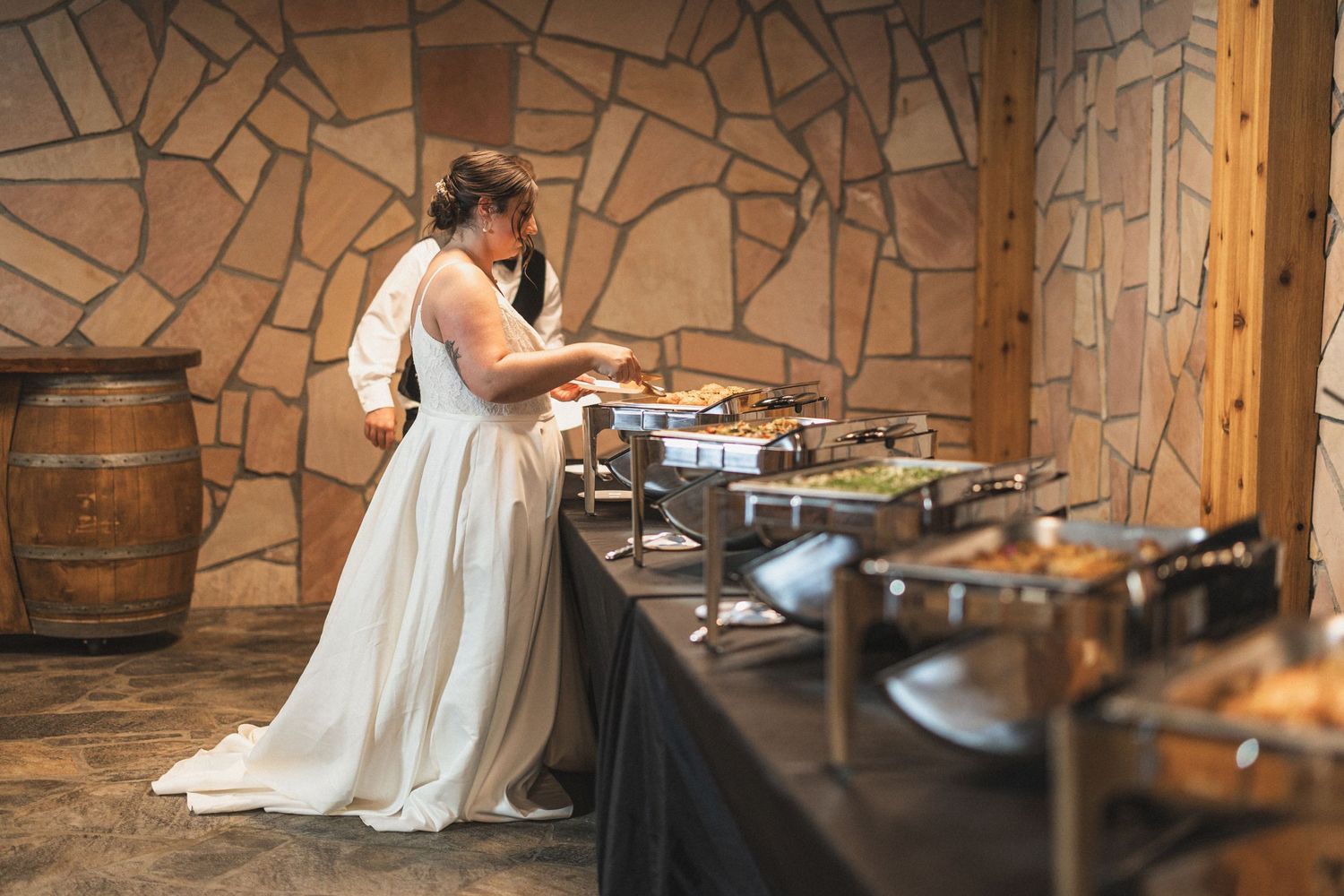 Bride in white gown serving food at wedding reception buffet with rustic stone wall background.