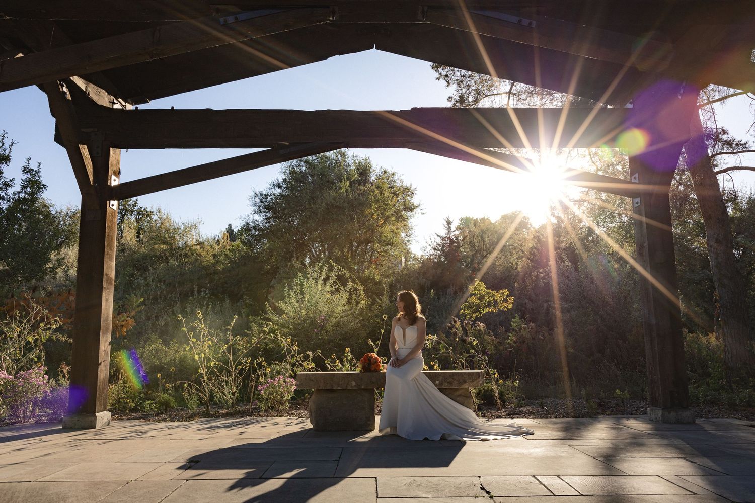 Bride in white dress poses under wooden pavilion during golden hour sunset.