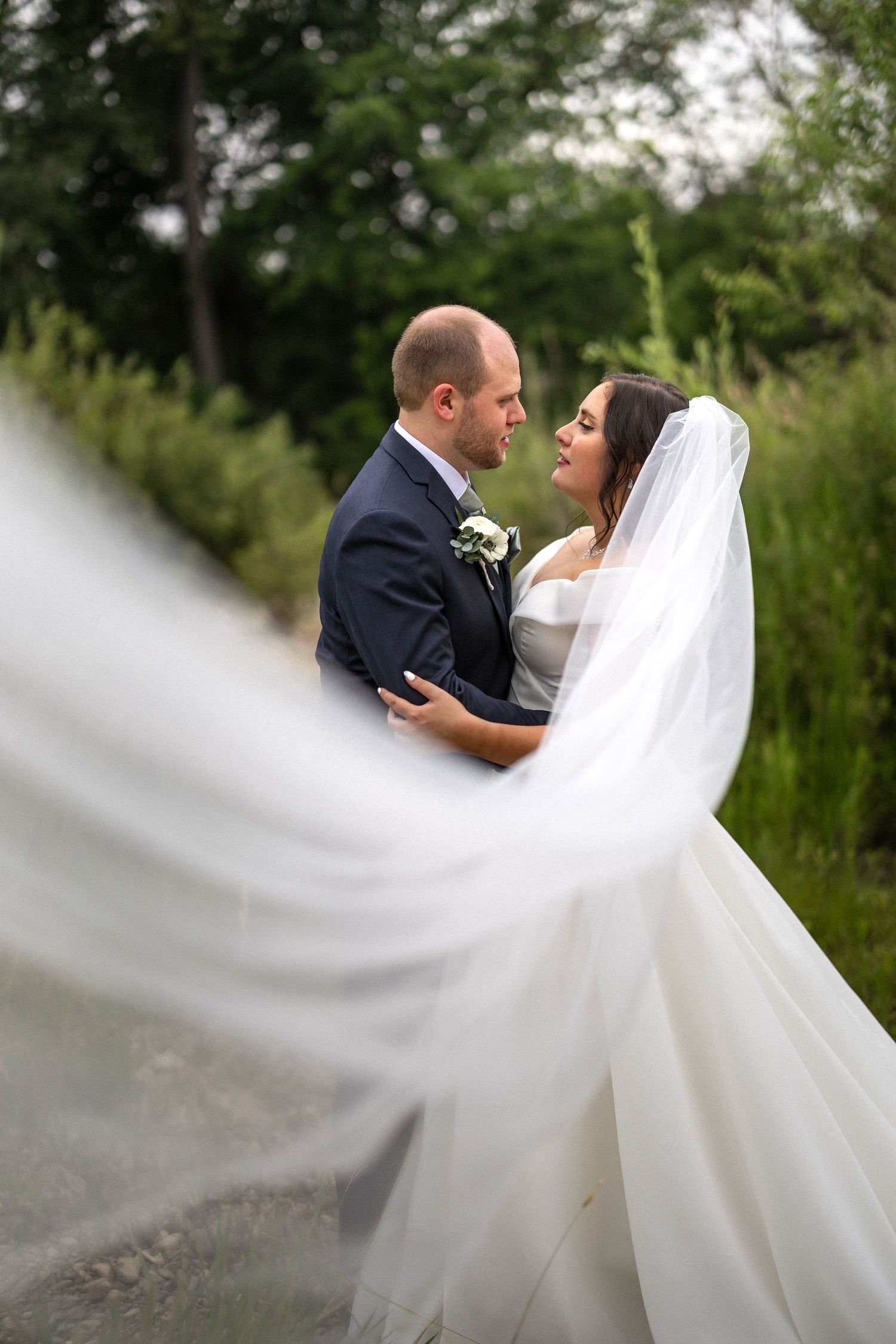 Bride in flowing white veil poses with partner against natural outdoor backdrop.