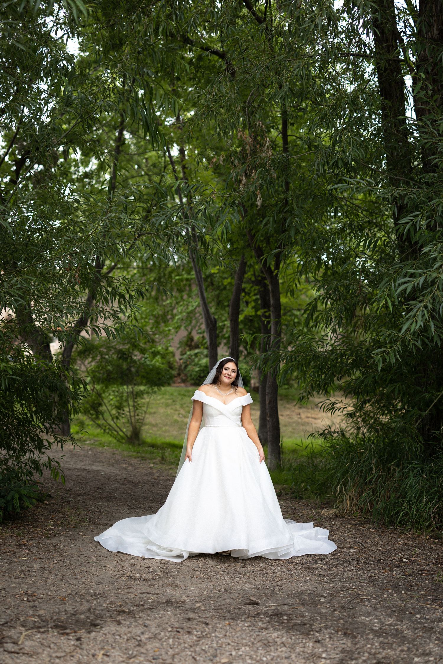 A bride in a voluminous white wedding dress sits elegantly on a natural dirt path surrounded by green foliage.