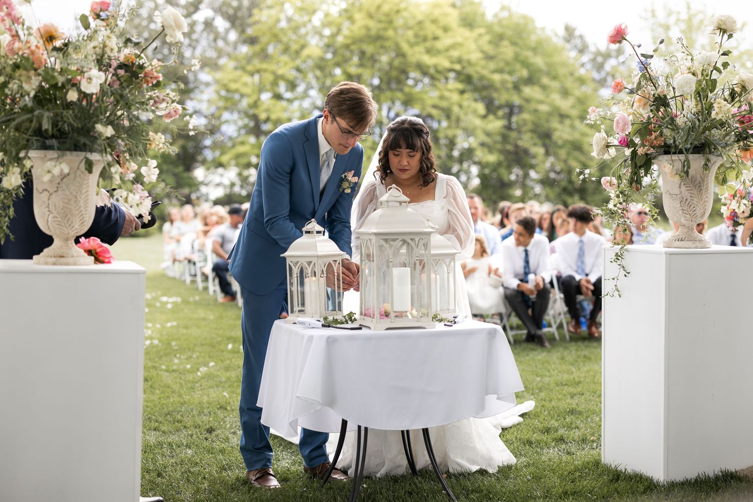 Couple in formal attire performing a unity ceremony with lanterns at an outdoor wedding.