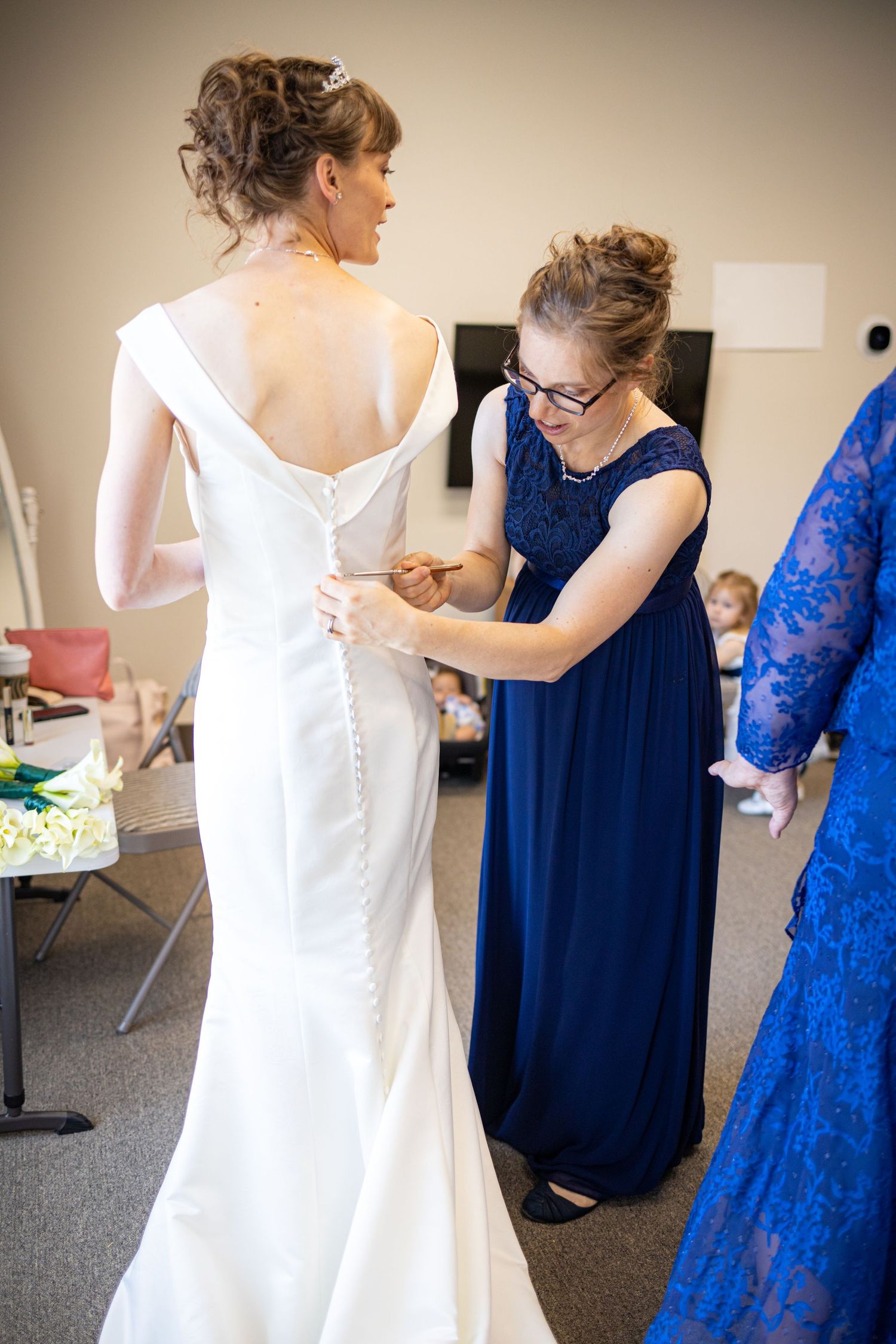 Wedding dress being buttoned up by attendant in navy blue dress during preparation.
