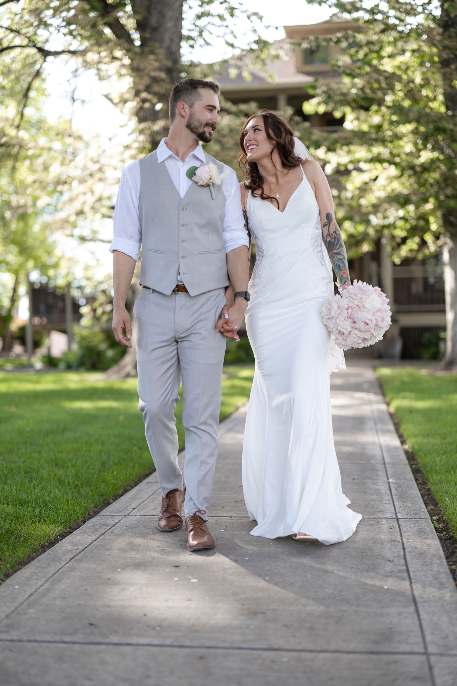 Elegant wedding couple walking together on a concrete path with trees and building in background.