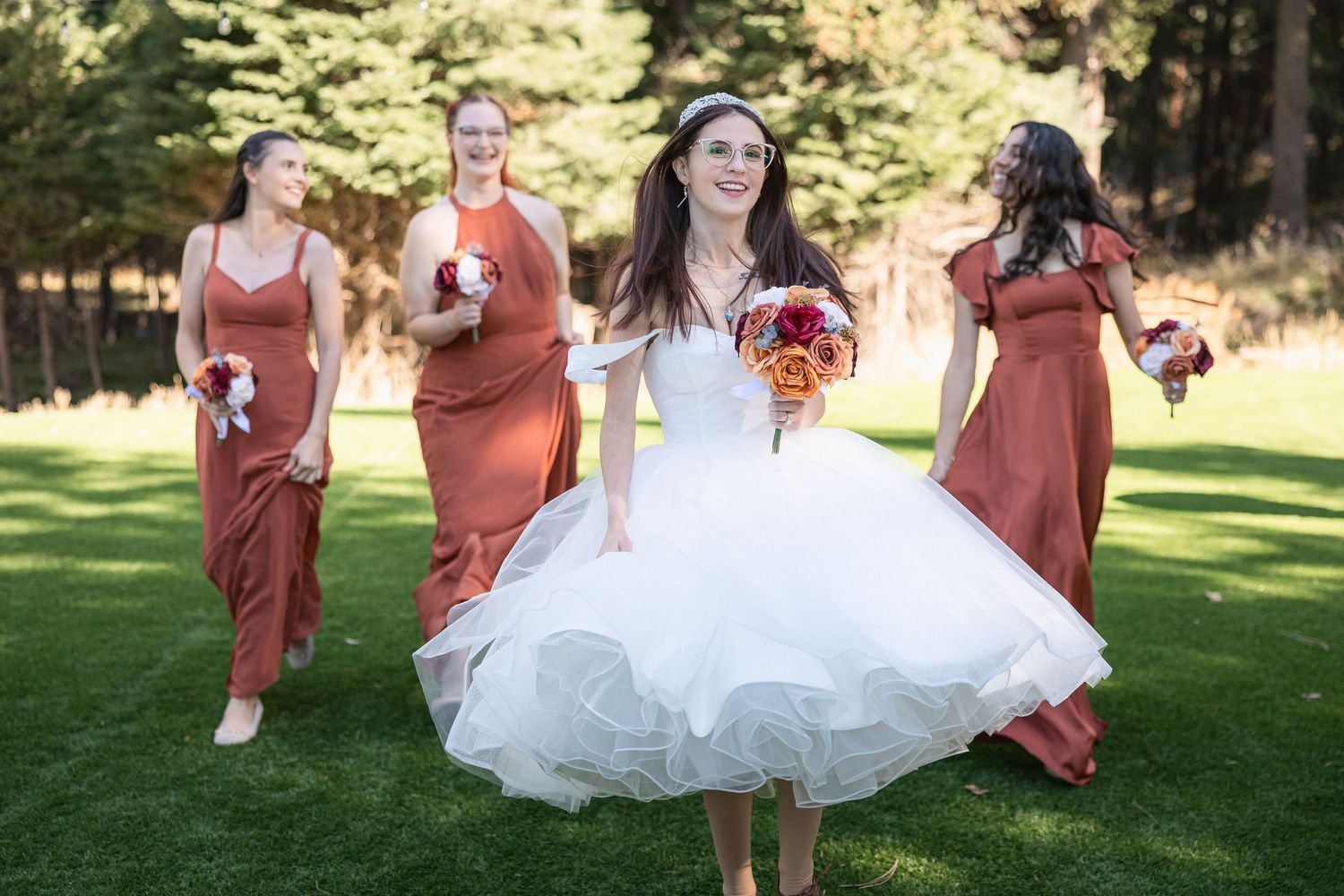 Joyful bridal party in terracotta dresses celebrates on sunny lawn with evergreens.