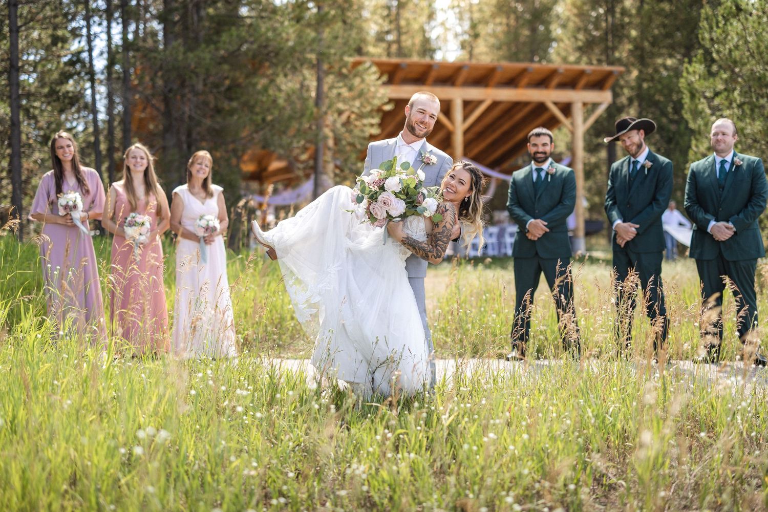 Wedding celebration group photo with bridal party and wooden pergola in natural landscape.