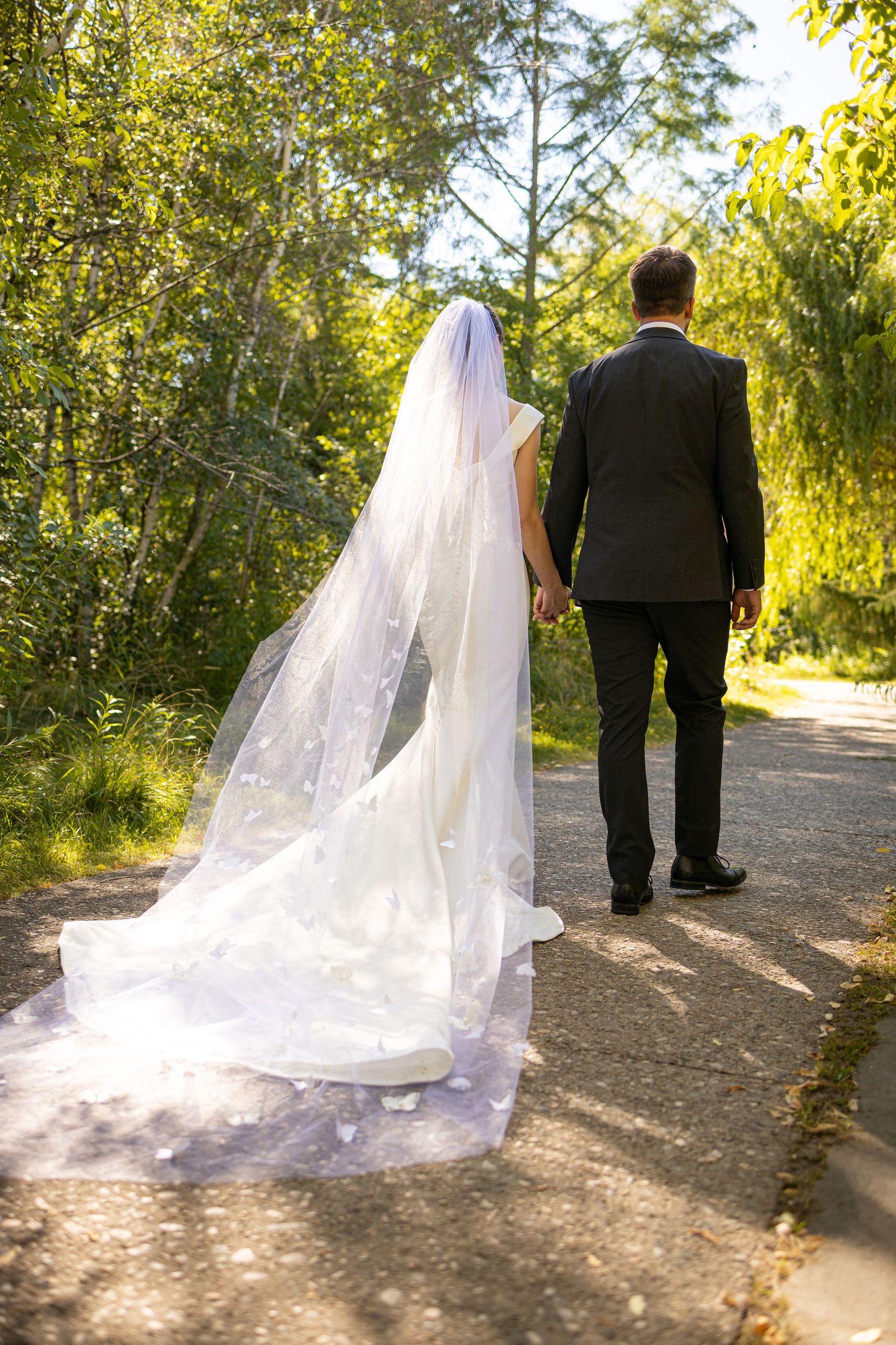 Bride and groom walking down a sunlit garden path with flowing white veil trailing behind.
