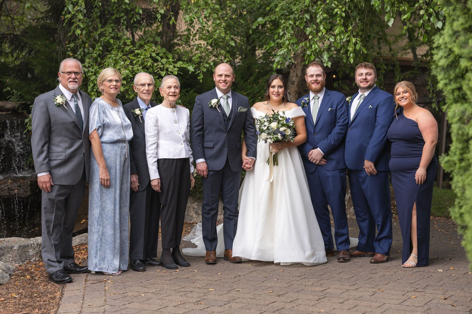 Formal wedding party group photo outdoors with brick wall and greenery backdrop.