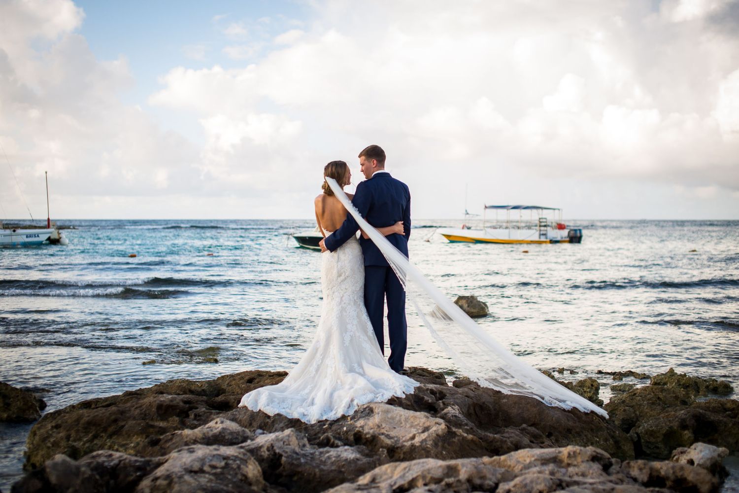 A couple shares a romantic moment on rocky coastal shores with boats and ocean waves in the background at sunset.