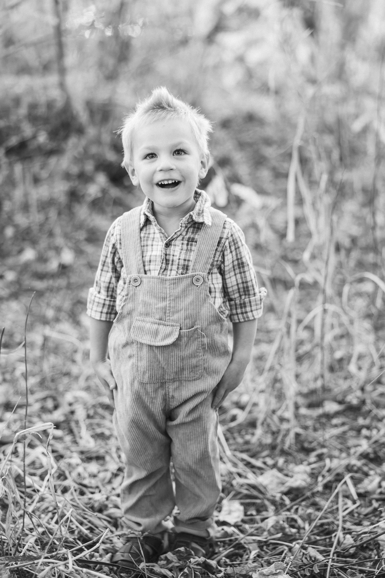 Black and white portrait of small child in overalls playing in tall grass.