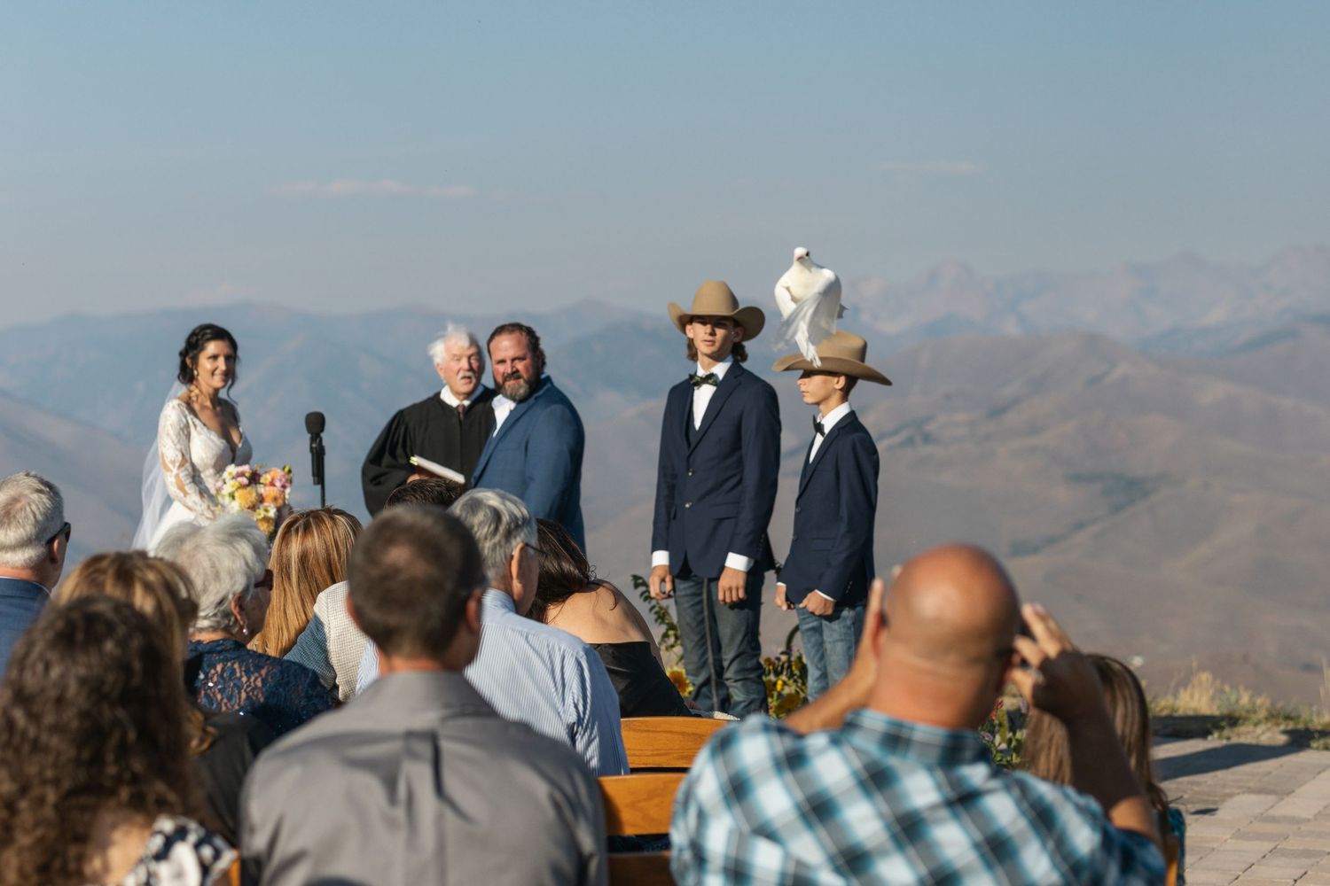 Outdoor mountain wedding ceremony with guests seated on wooden benches overlooking dramatic landscape.