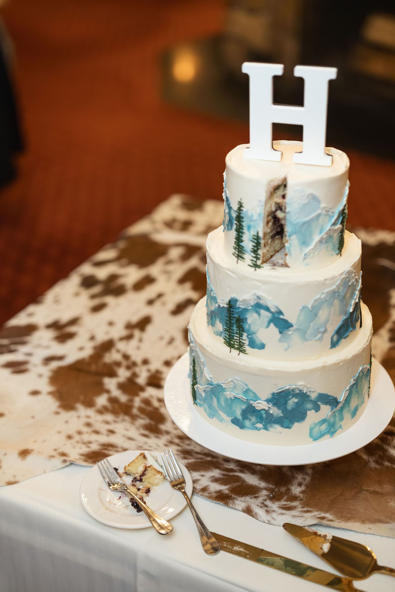 Three-tiered white wedding cake decorated with blue watercolor mountains and pine trees topped with letter H.