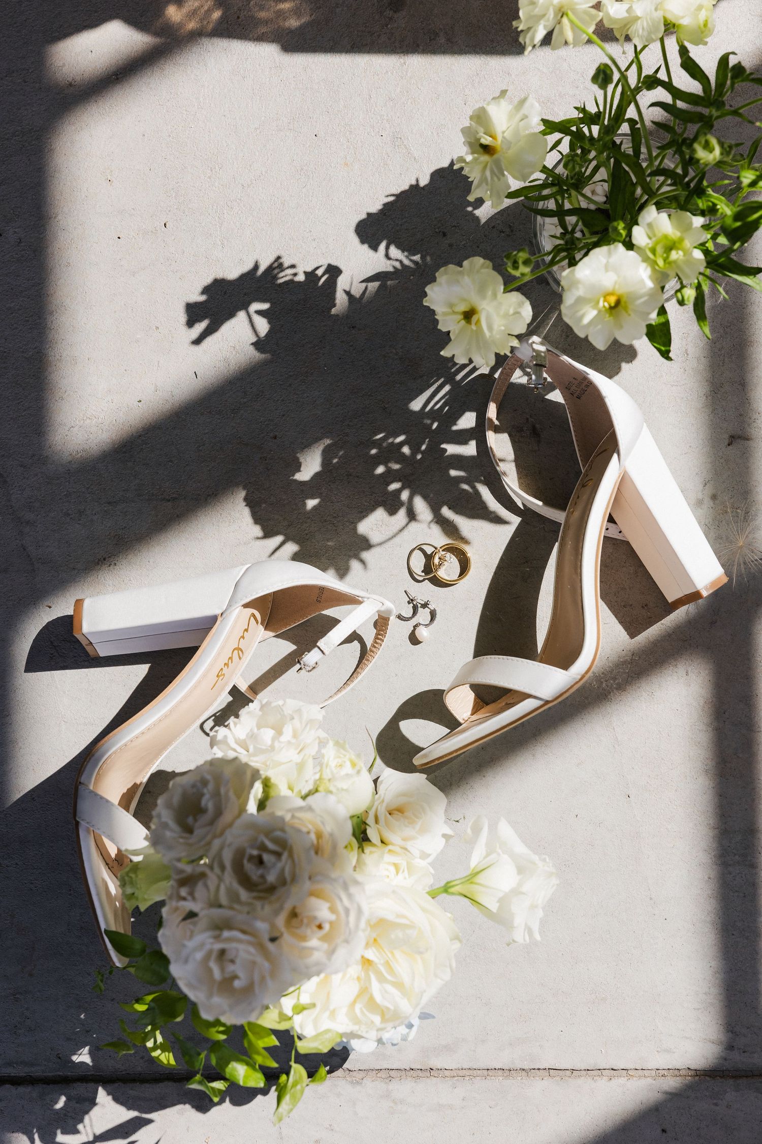 White bridal heels with gold wedding rings and white roses bouquet arranged on concrete in sunlight.