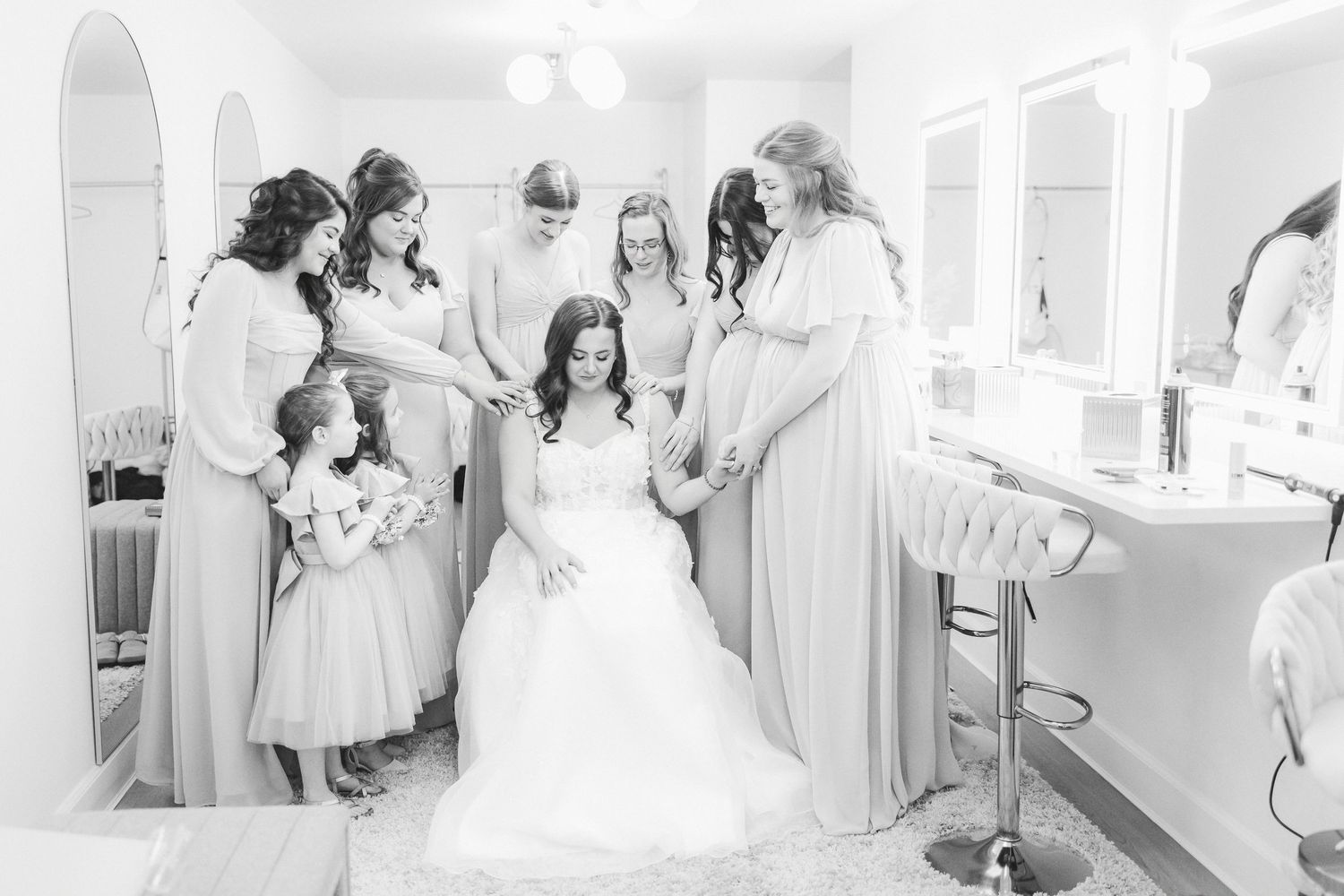 Bride seated in wedding gown surrounded by bridesmaids and flower girls in a bridal suite, black and white photo.