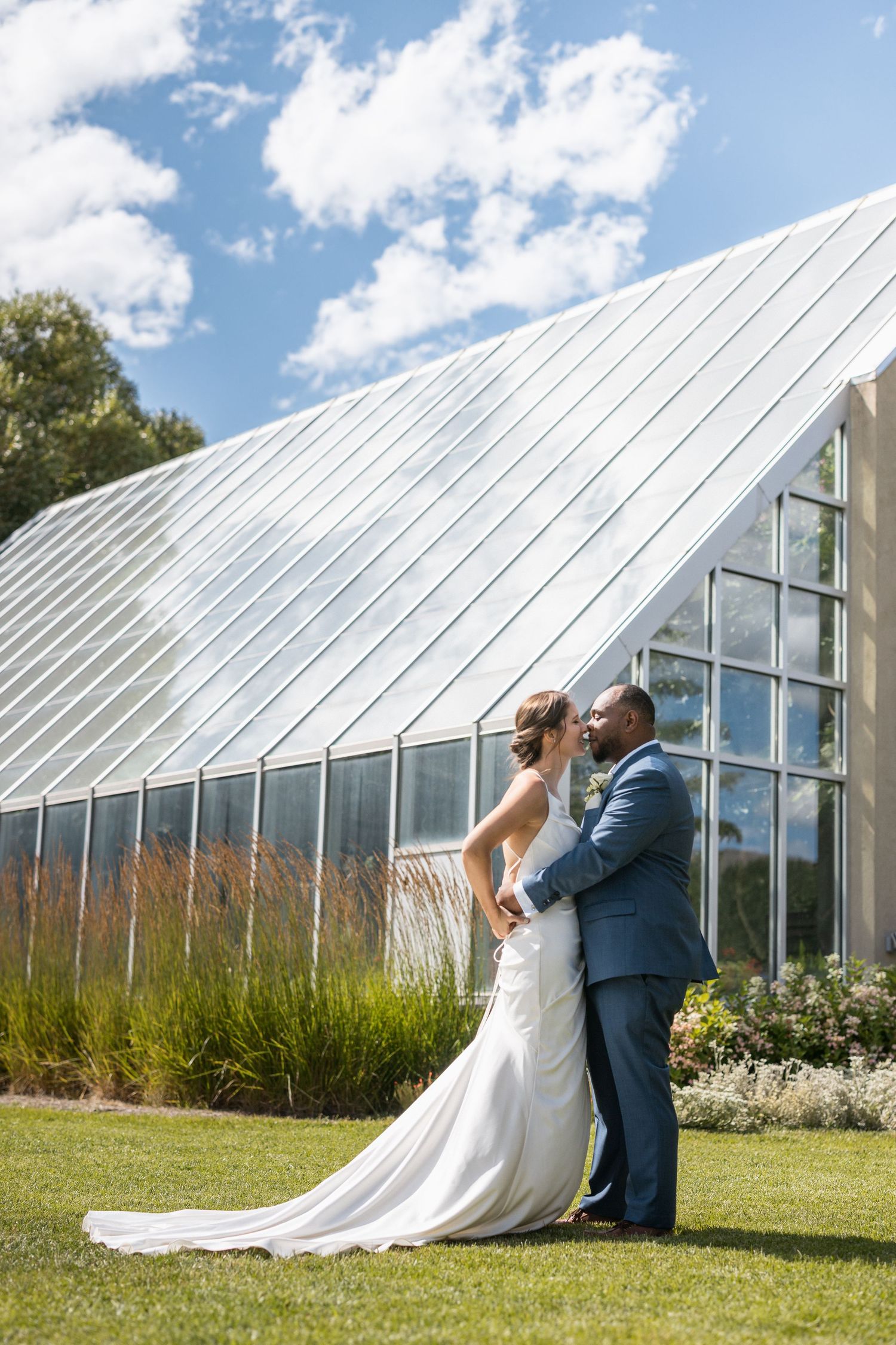 Wedding couple poses playfully in front of large greenhouse structures under blue skies.
