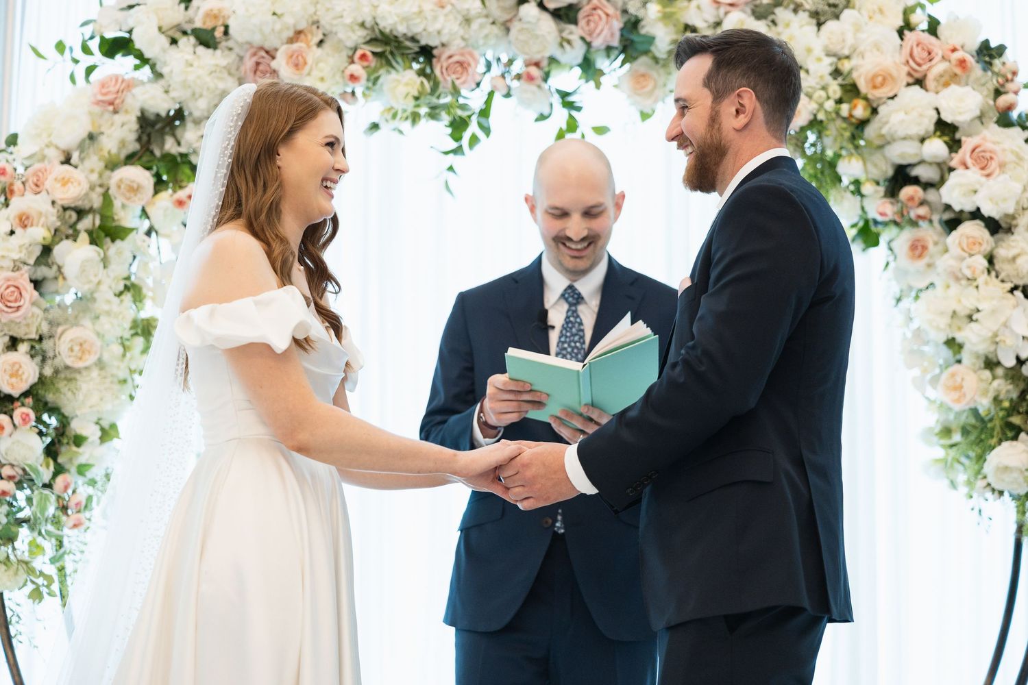 Wedding ceremony in progress under an elegant floral arch with pink and white roses and greenery.
