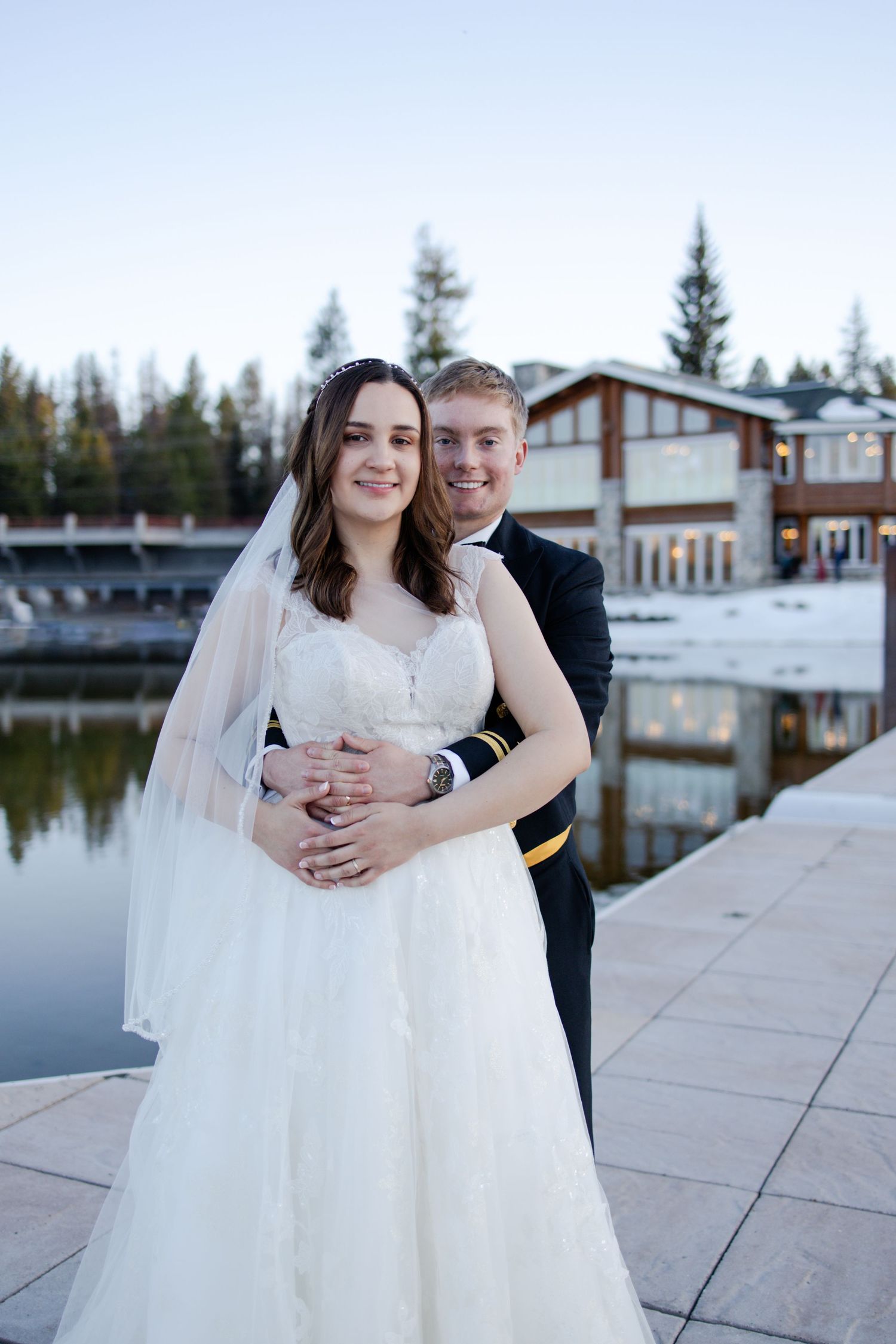 A wedding couple poses by a serene lake with a modern lodge in the background during golden hour.