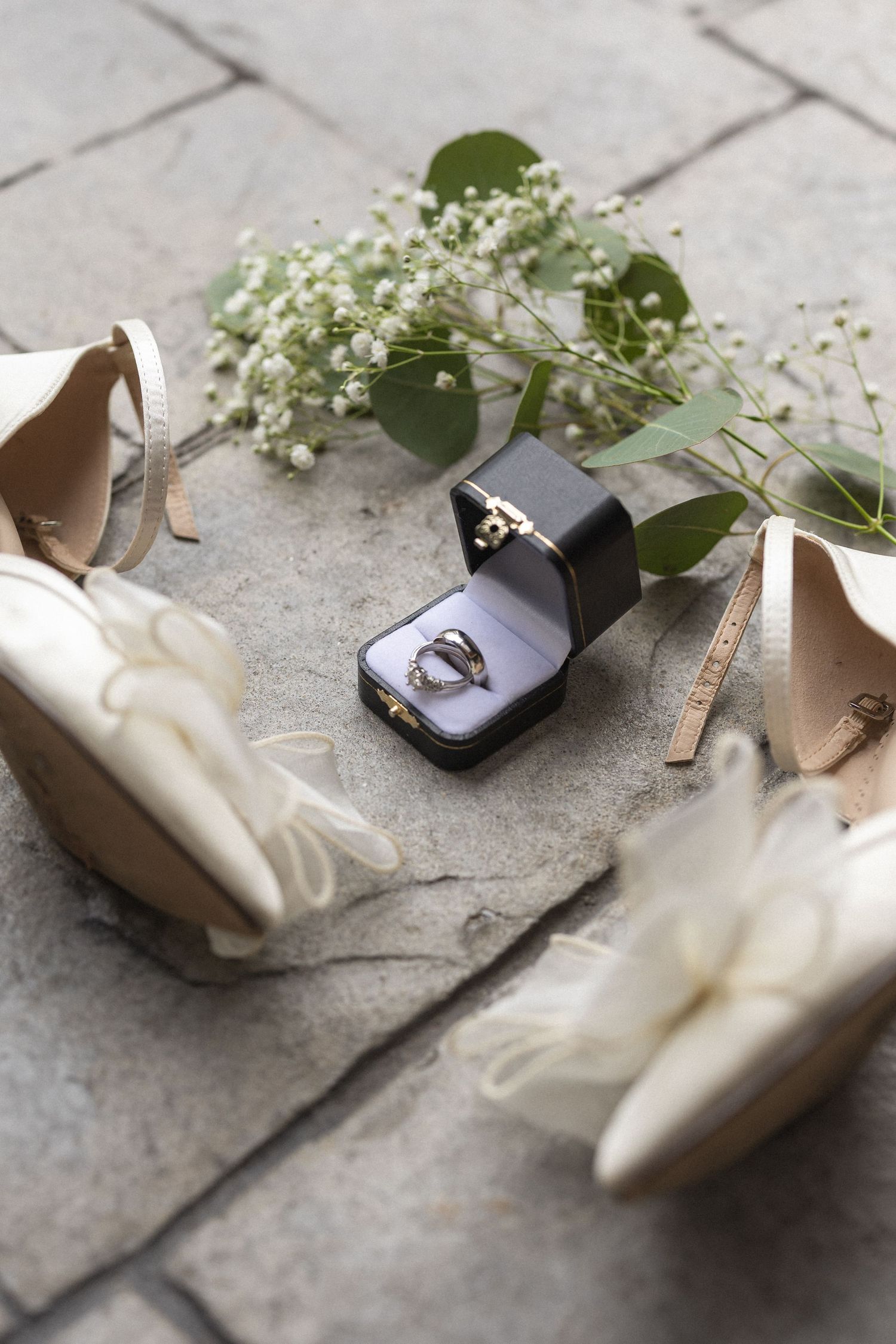 Wedding details flat lay with white heels, rings, and baby's breath flowers on stone.