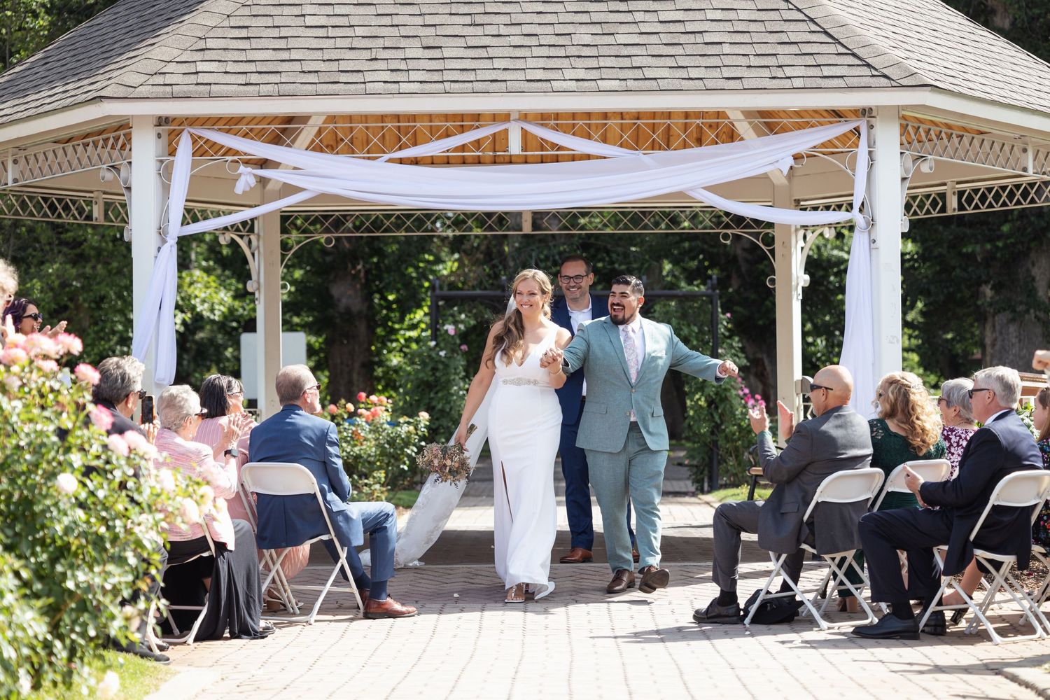 Couple exits wedding ceremony under white gazebo while guests applaud.