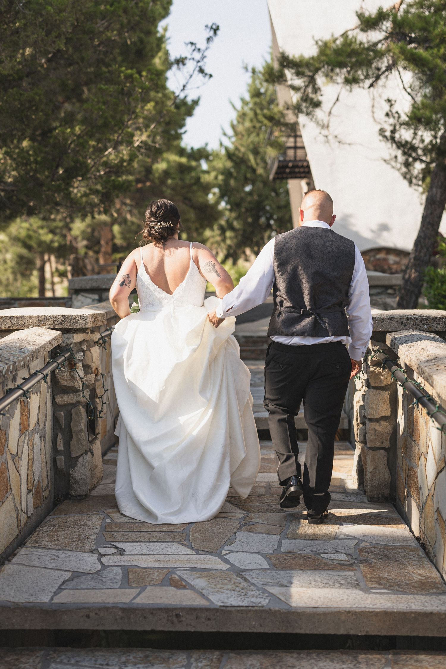A couple walks up stone steps together in their wedding attire surrounded by pine trees.