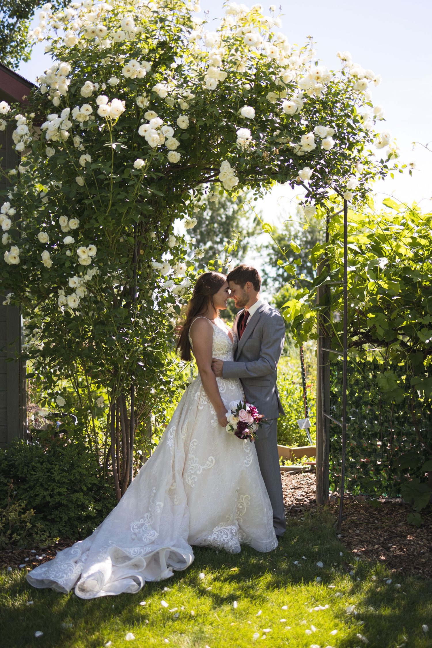 Newlyweds embrace under a white rose arbor in a romantic garden setting.