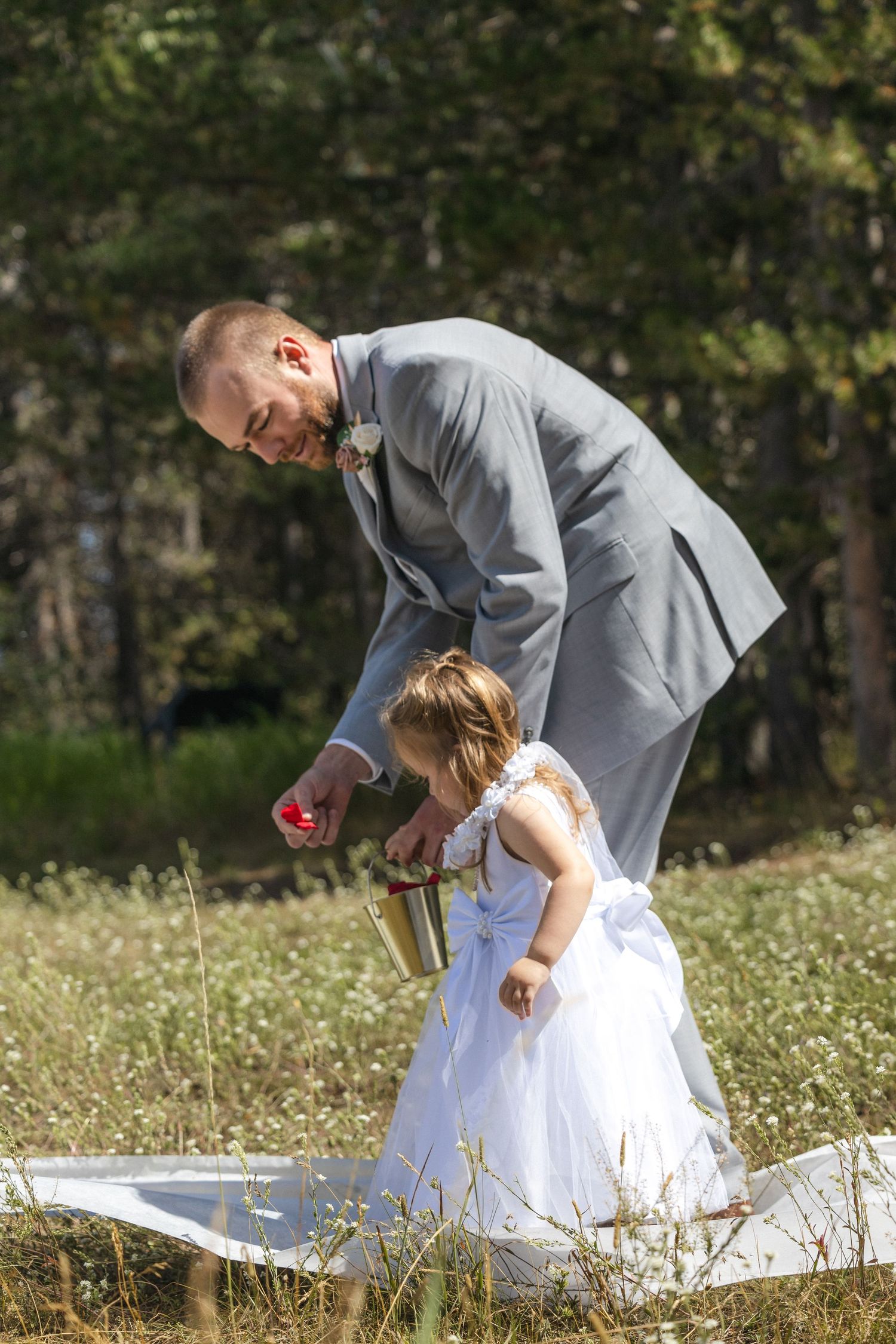 A person in grey suit kneeling next to small child in white dress outdoors.