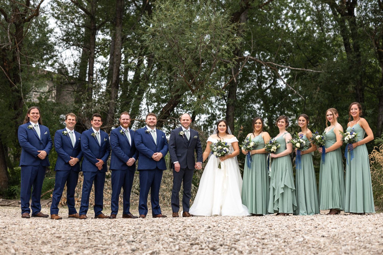 A large wedding party in navy suits and sage green dresses poses together on a wooded path.