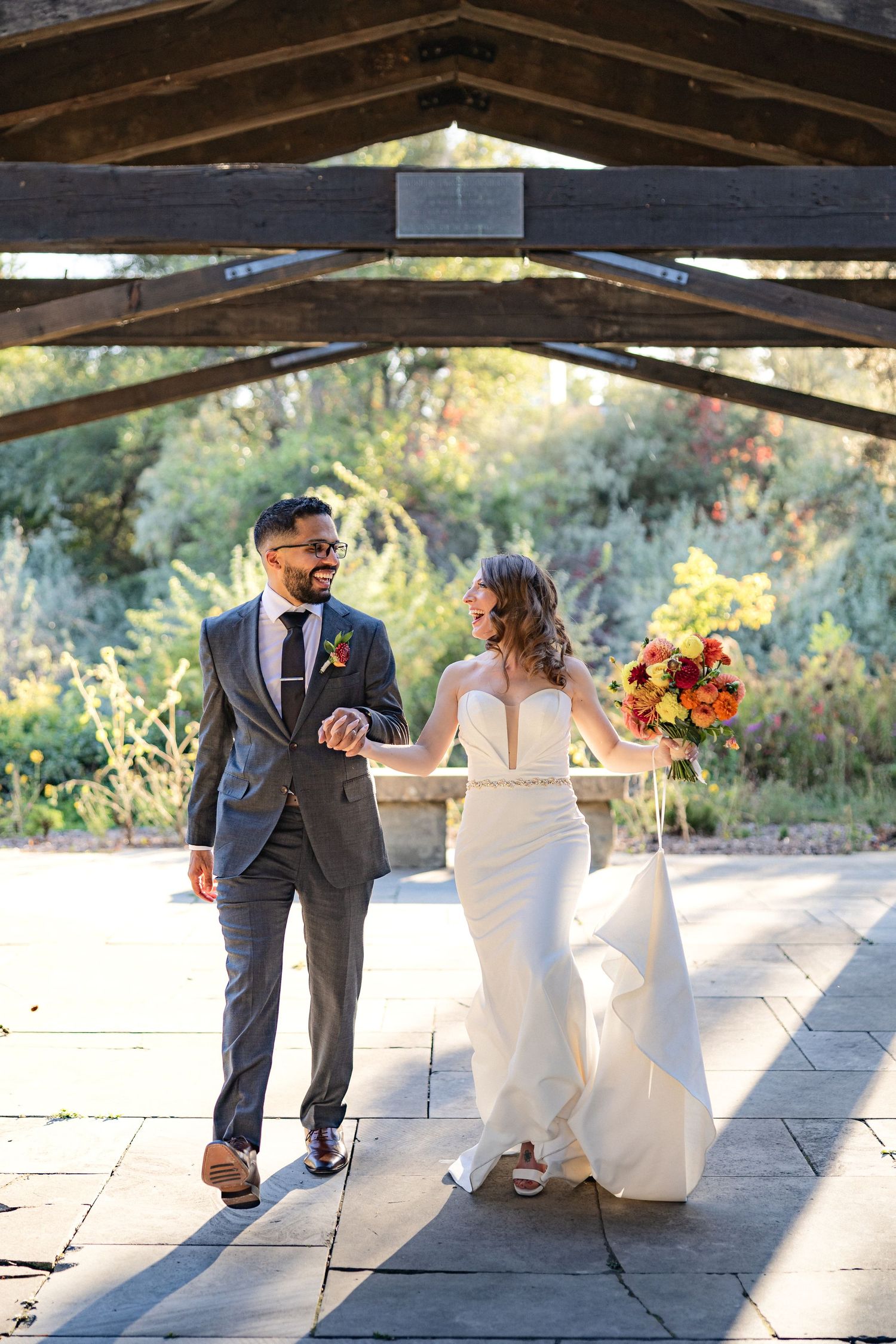 Happy couple walks together on stone path under rustic wooden pergola after ceremony.