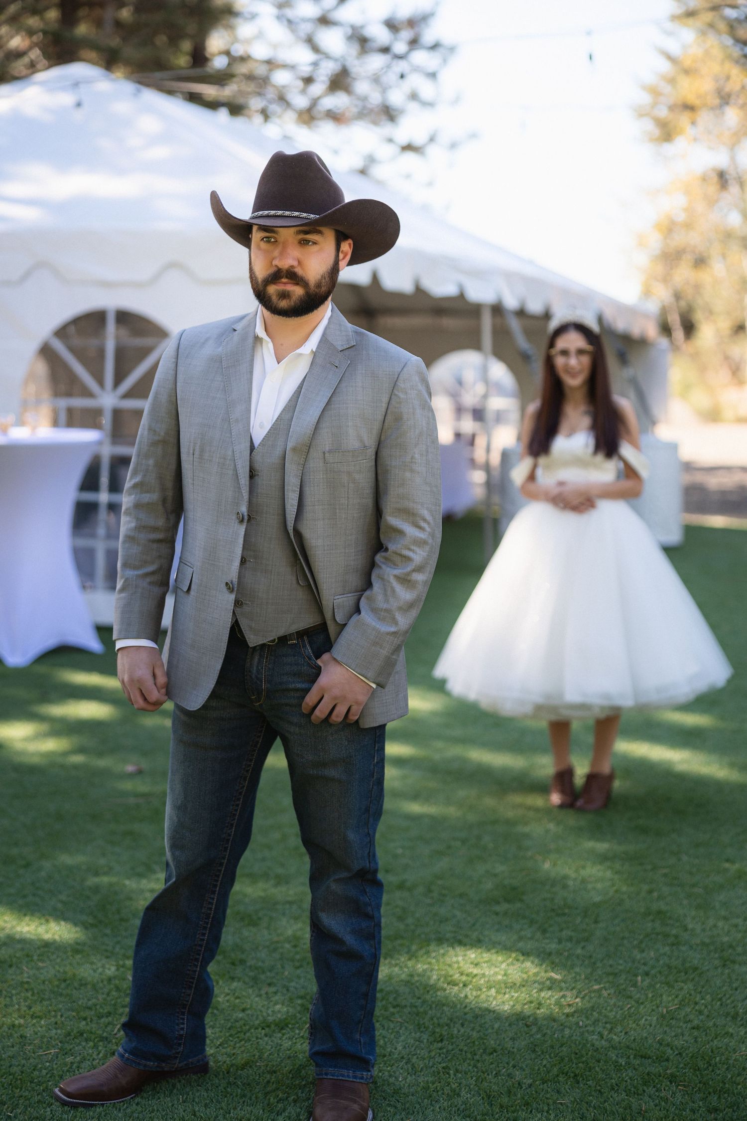 A western-themed wedding photo showing a groomsman in cowboy attire with a bride in the background by a white tent.