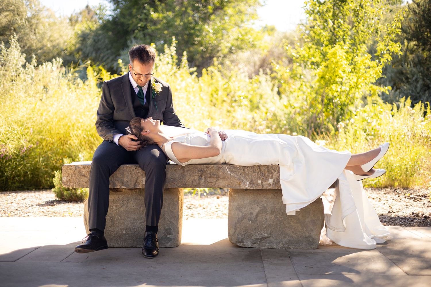 Newlyweds pose playfully on stone bench with flowing white dress in sunlit garden.