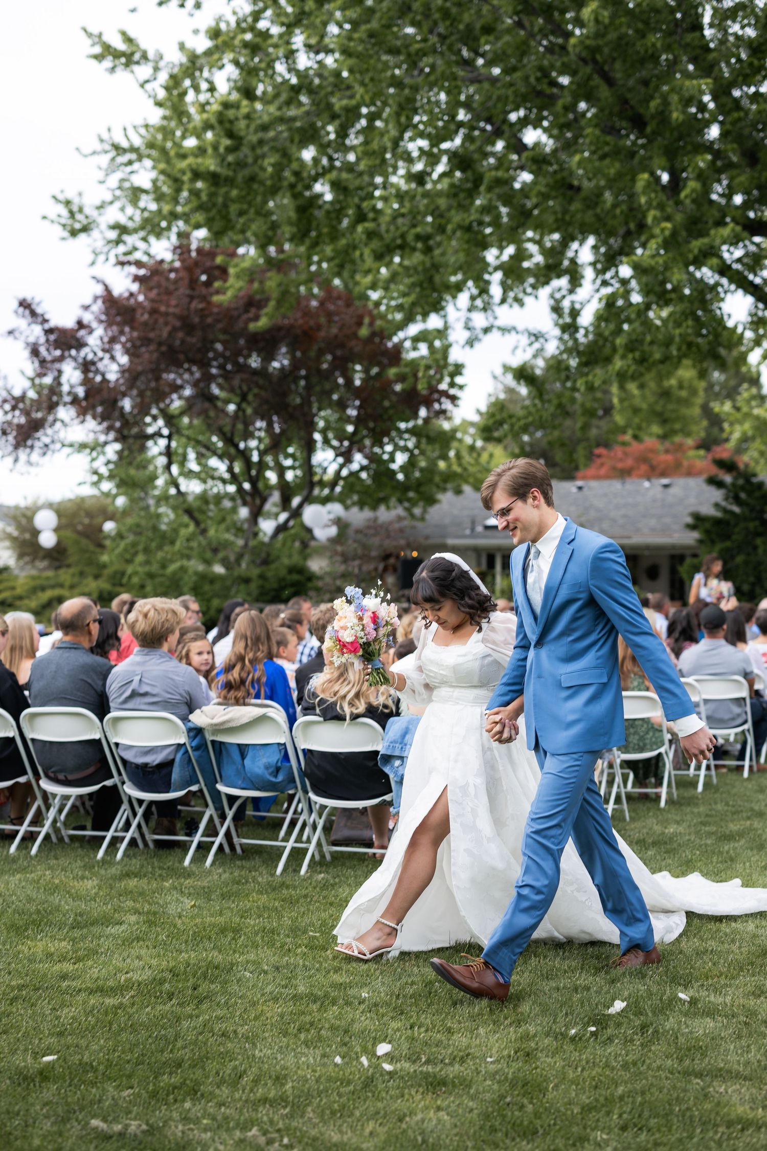 Couple walking together on grass between rows of folding chairs at outdoor wedding ceremony.